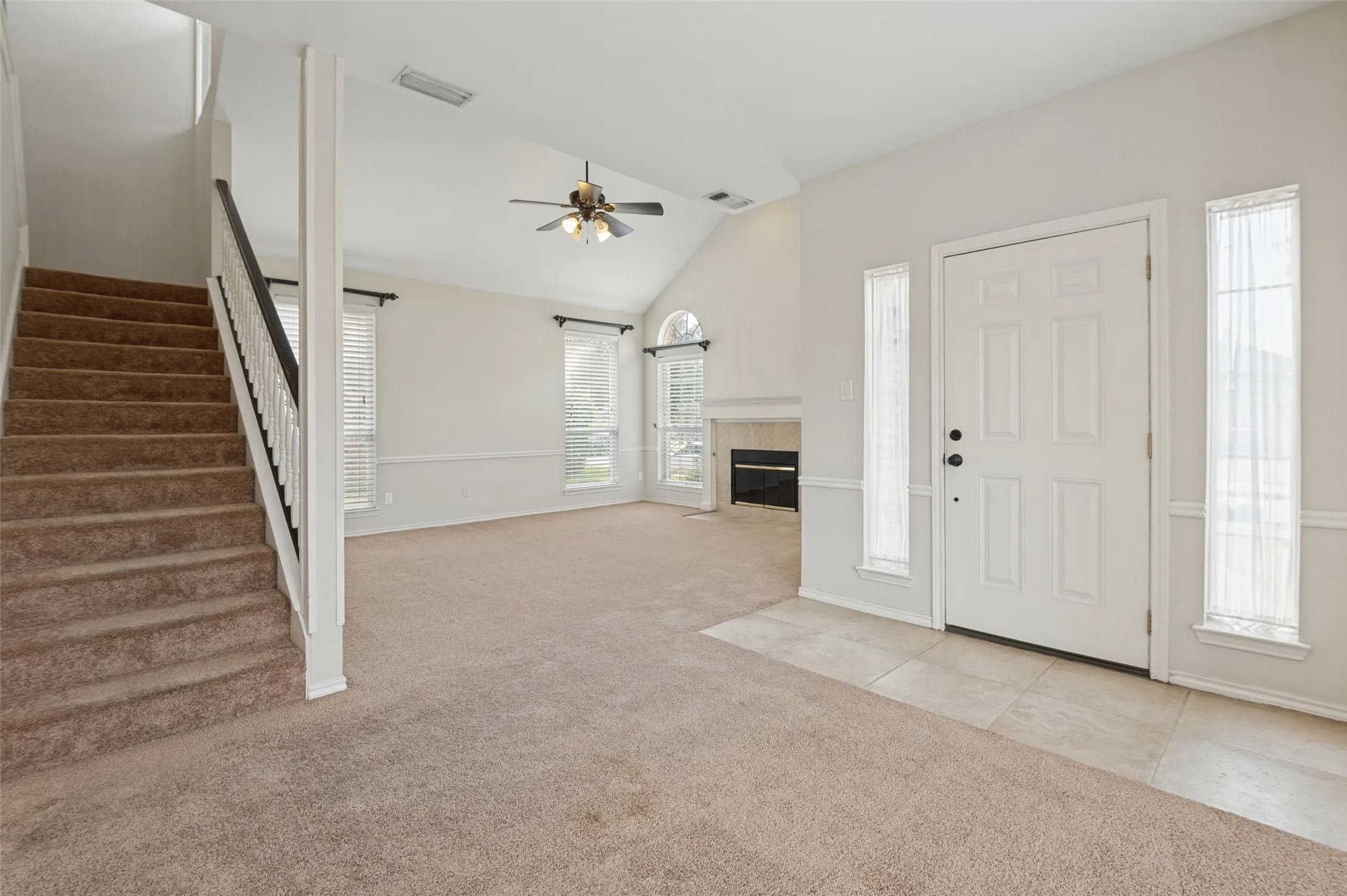 Vaulted ceiling with ceiling fan, white paneled entry door with sidelights, staircase with dark wood-finish handrail, fireplace with mantel, and carpeted flooring