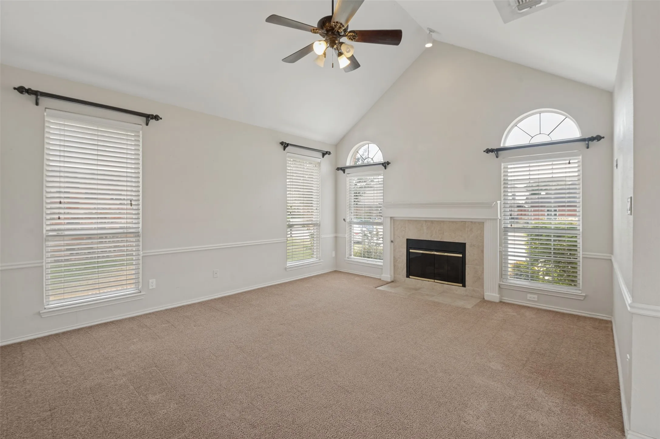 Vaulted ceiling with an attached ceiling fan, floor-to-ceiling fireplace with stone surround, arched transom windows, and neutral carpeting