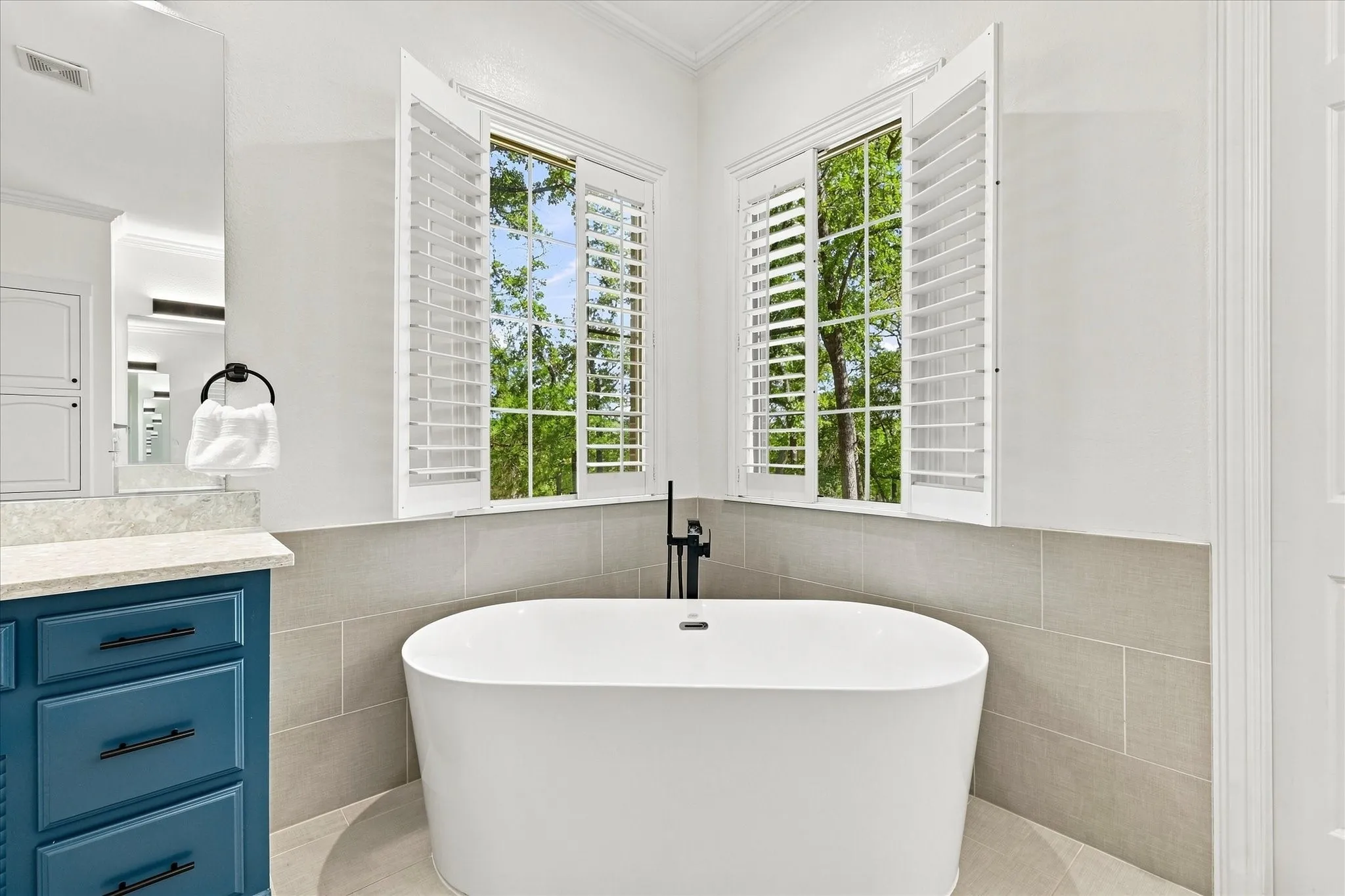 Freestanding soaking tub with a floor-mounted faucet, plantation shutters, tiled wainscoting, and a vanity with a countertop