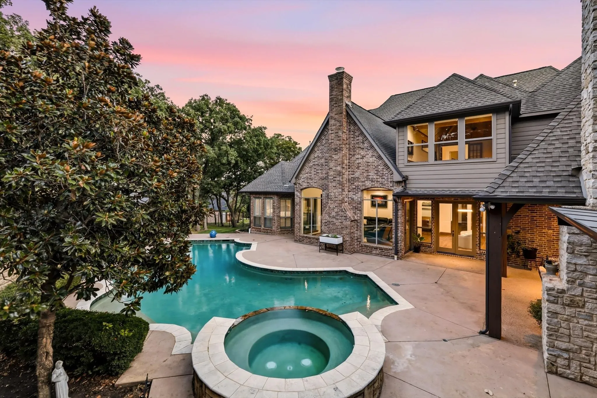 Curved swimming pool with an integrated spa, extensive concrete patio, multi-level home exterior with brick and stone accents, and a prominent brick chimney