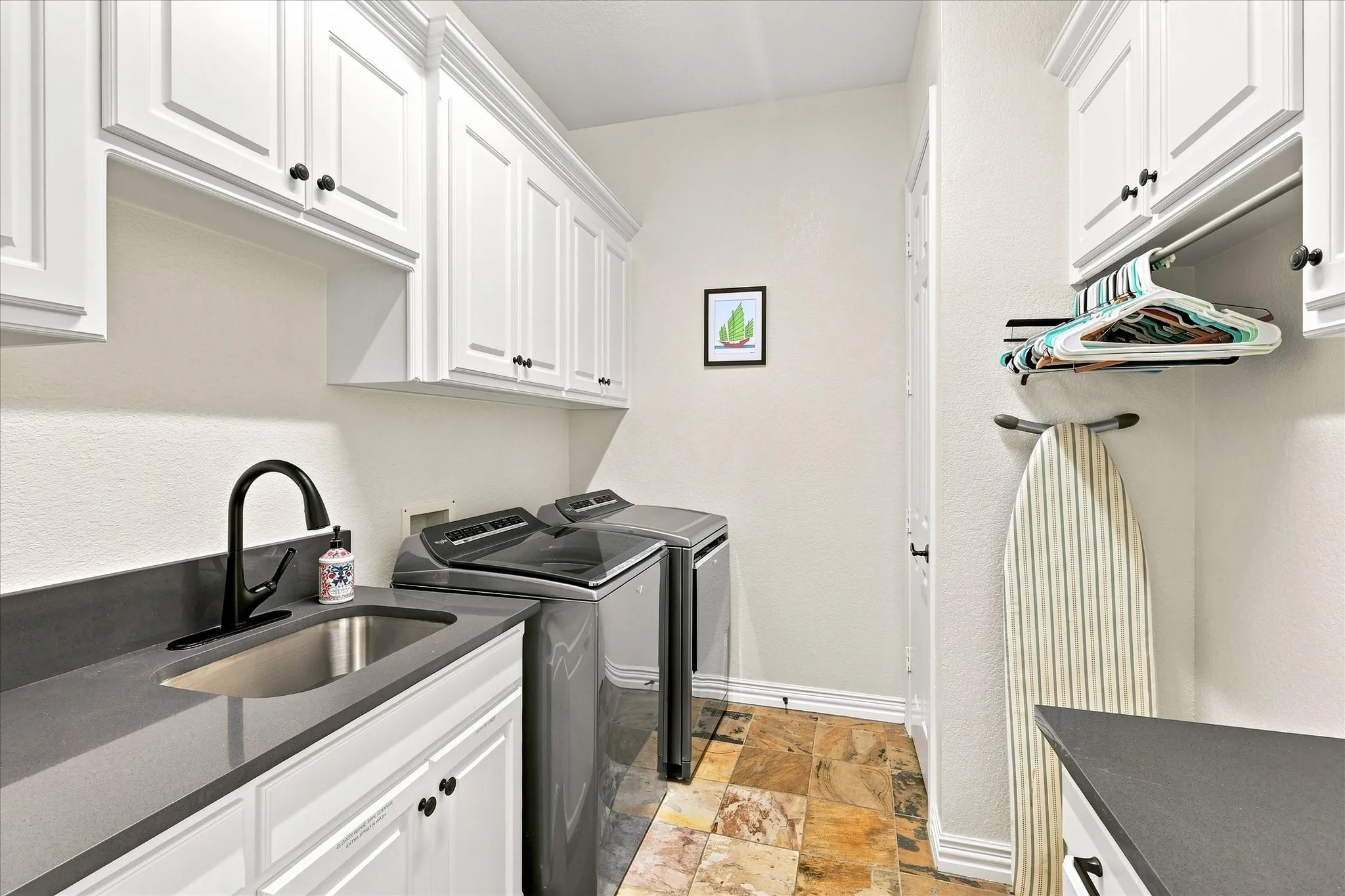 White shaker cabinetry, dark countertops, undermount sink with a matte black faucet, and stone-finish flooring