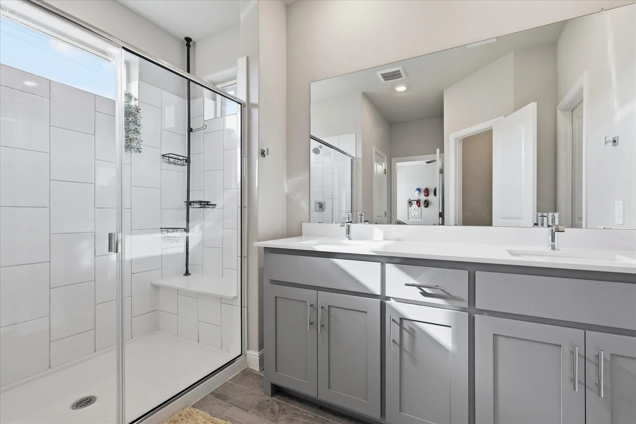 Shower enclosure with clear glass doors and a built-in bench, accompanied by a dual vanity featuring light gray cabinetry and integrated sinks