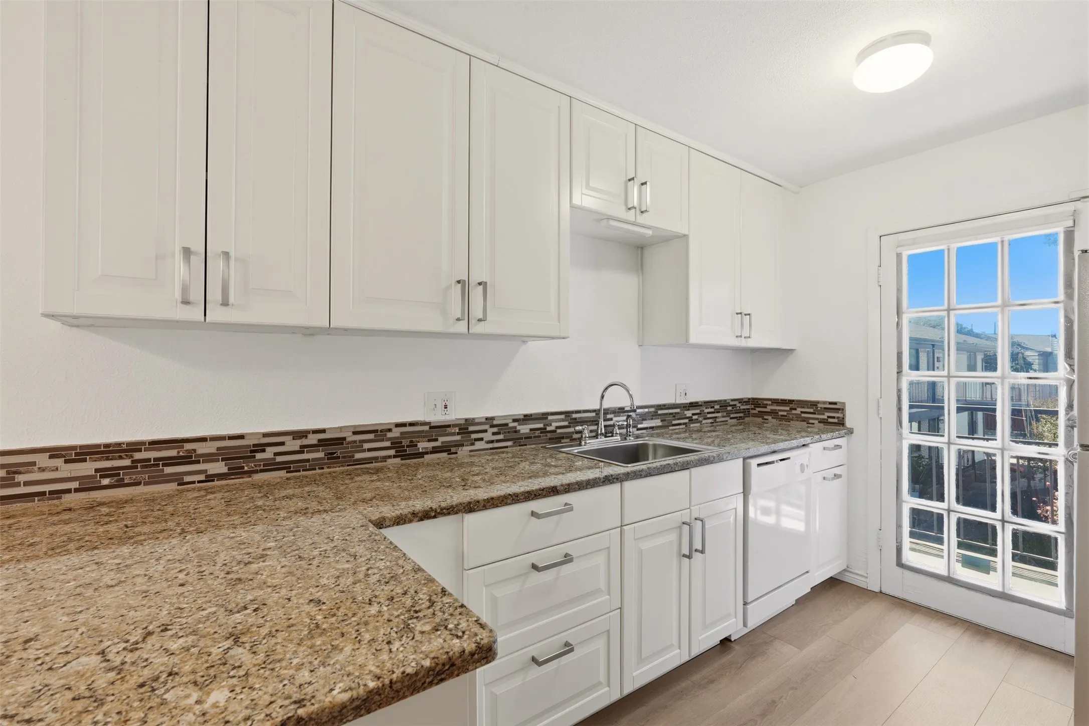 White cabinetry with brushed nickel hardware, granite-style countertops, mosaic tile backsplash, stainless steel sink, and wood-finish flooring