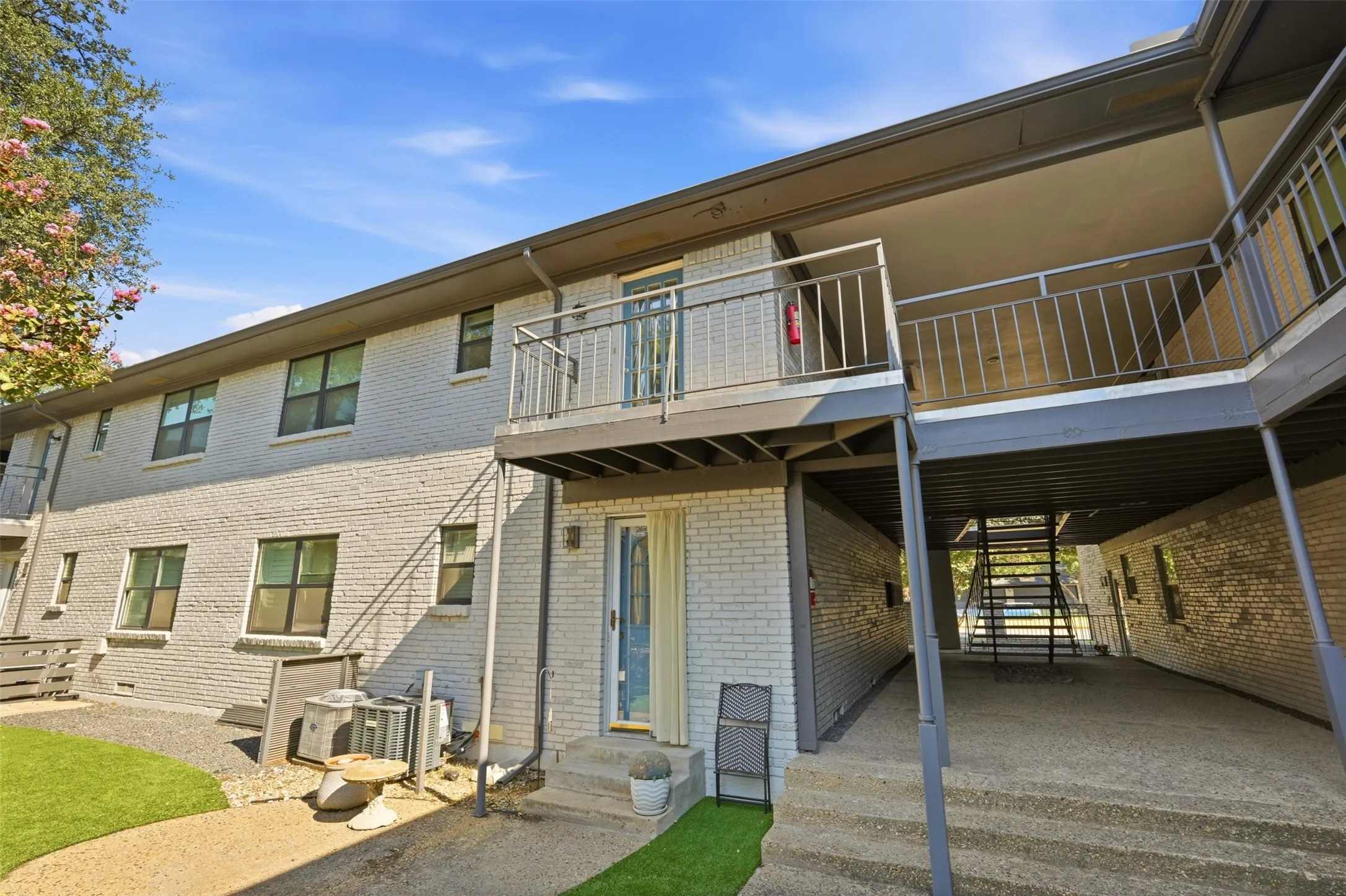 Two-story exterior with light-colored brick facade, multiple balconies with metal railings, and a lower-level concrete patio
