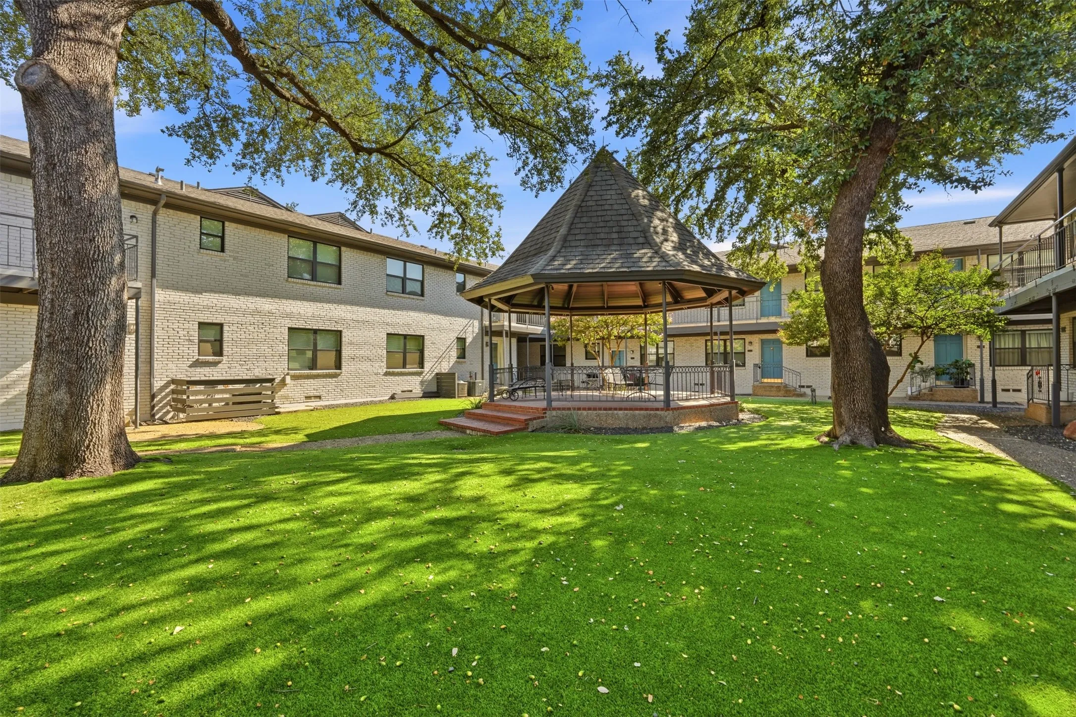Lush green lawn with mature trees, a central gazebo with a shingled roof, and brick-clad buildings