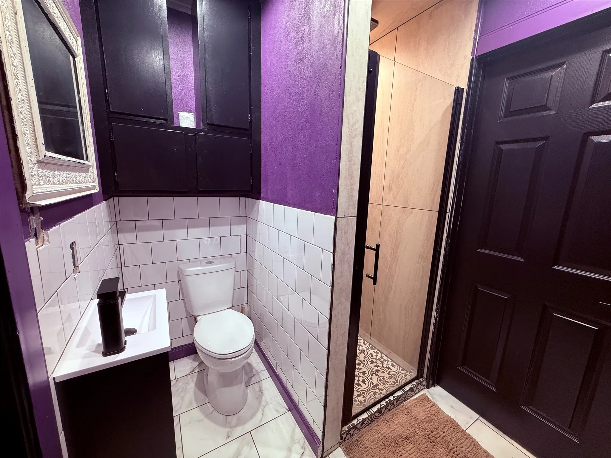 Compact bathroom featuring a white subway tile wainscoting, a white vanity with an integrated sink, a dark shower enclosure, and a paneled entry door