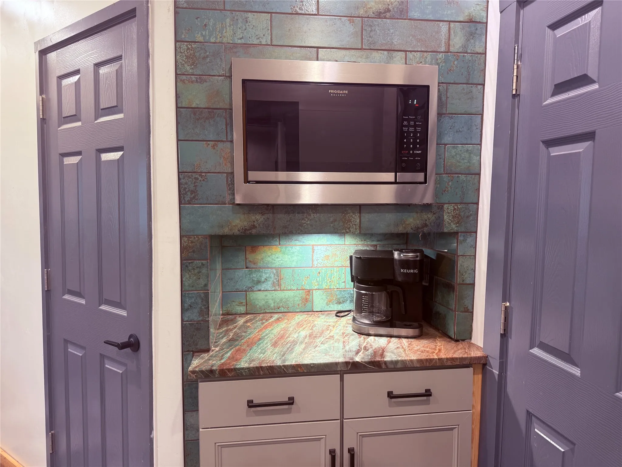 Built-in stainless steel microwave, unique variegated tile backsplash, under-cabinet lighting, gray cabinetry with dark hardware, and a colorful stone-finish countertop