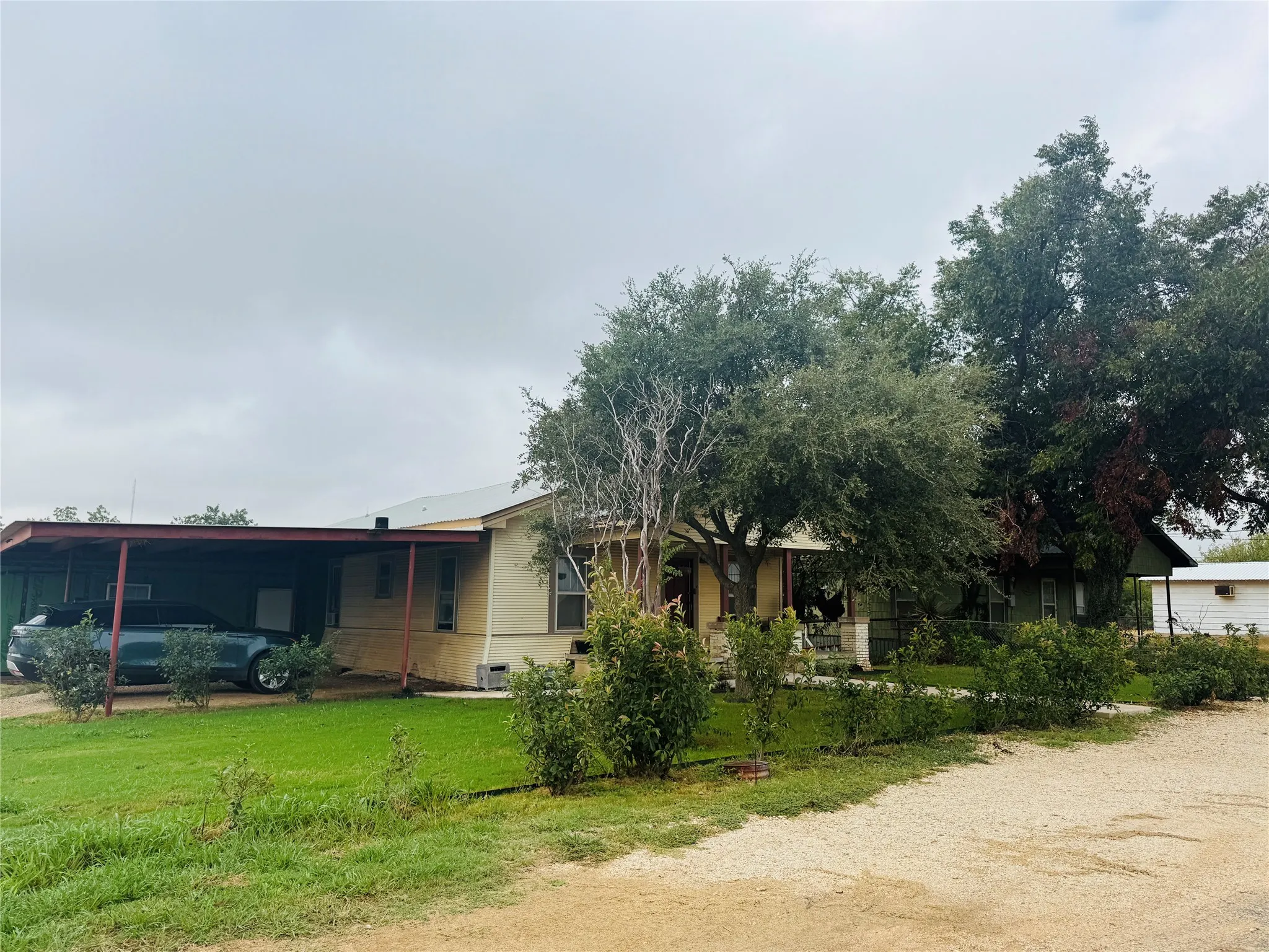 Carport with metal roof, established trees, perimeter landscaping, gravel driveway, and a manicured lawn