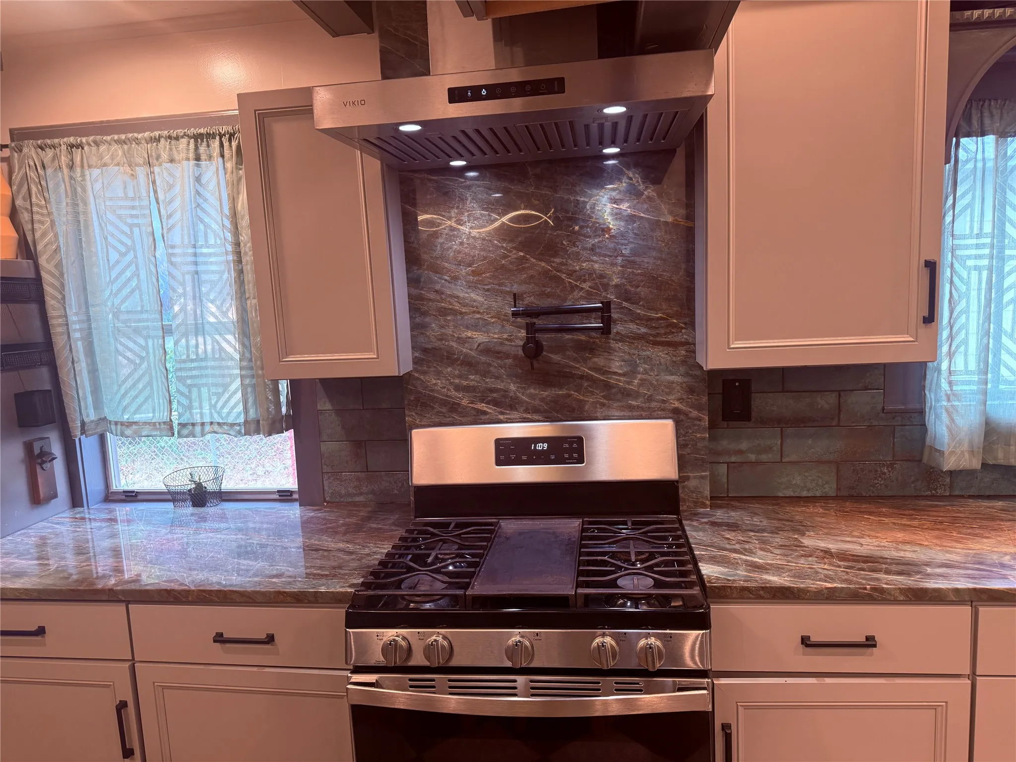 Stainless steel range with a pot filler, coordinating stainless steel range hood, stone countertops, and light-colored cabinetry with dark hardware