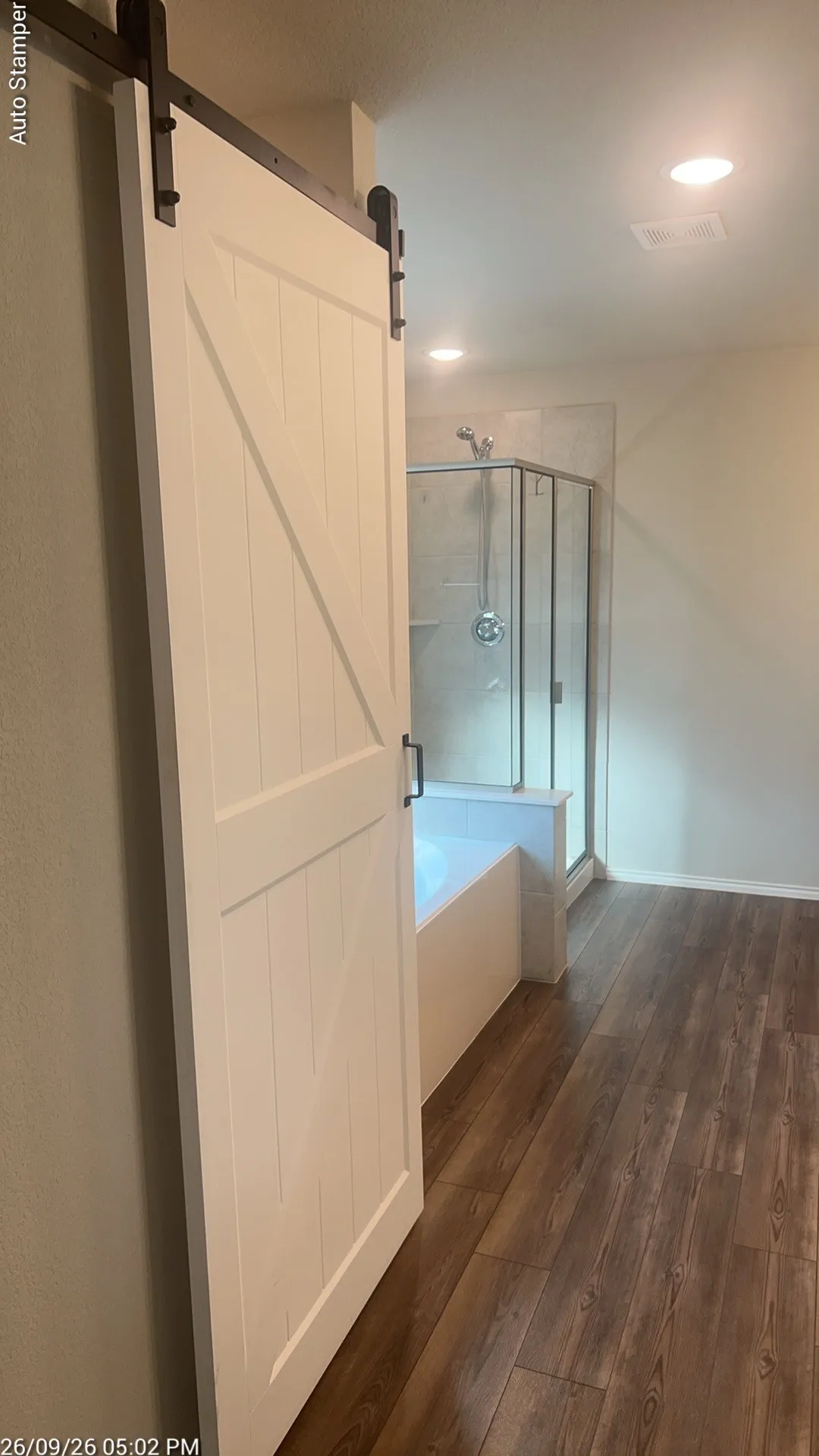 Wood-finish flooring throughout, white barn door with black hardware, built-in bathtub, and walk-in shower with glass enclosure