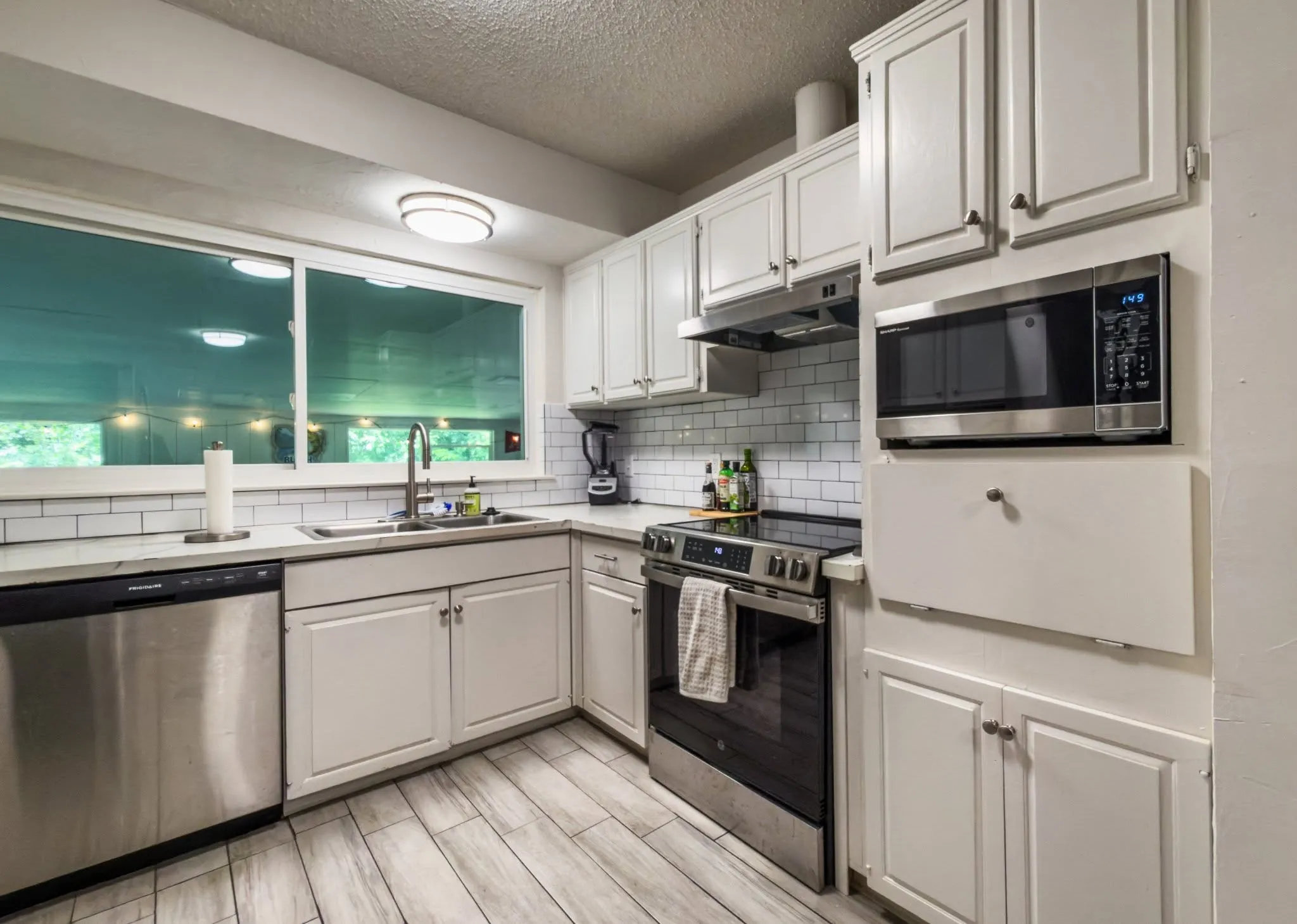 White cabinetry, stainless steel appliances, subway tile backsplash, light-toned wood-finish flooring, and a large window