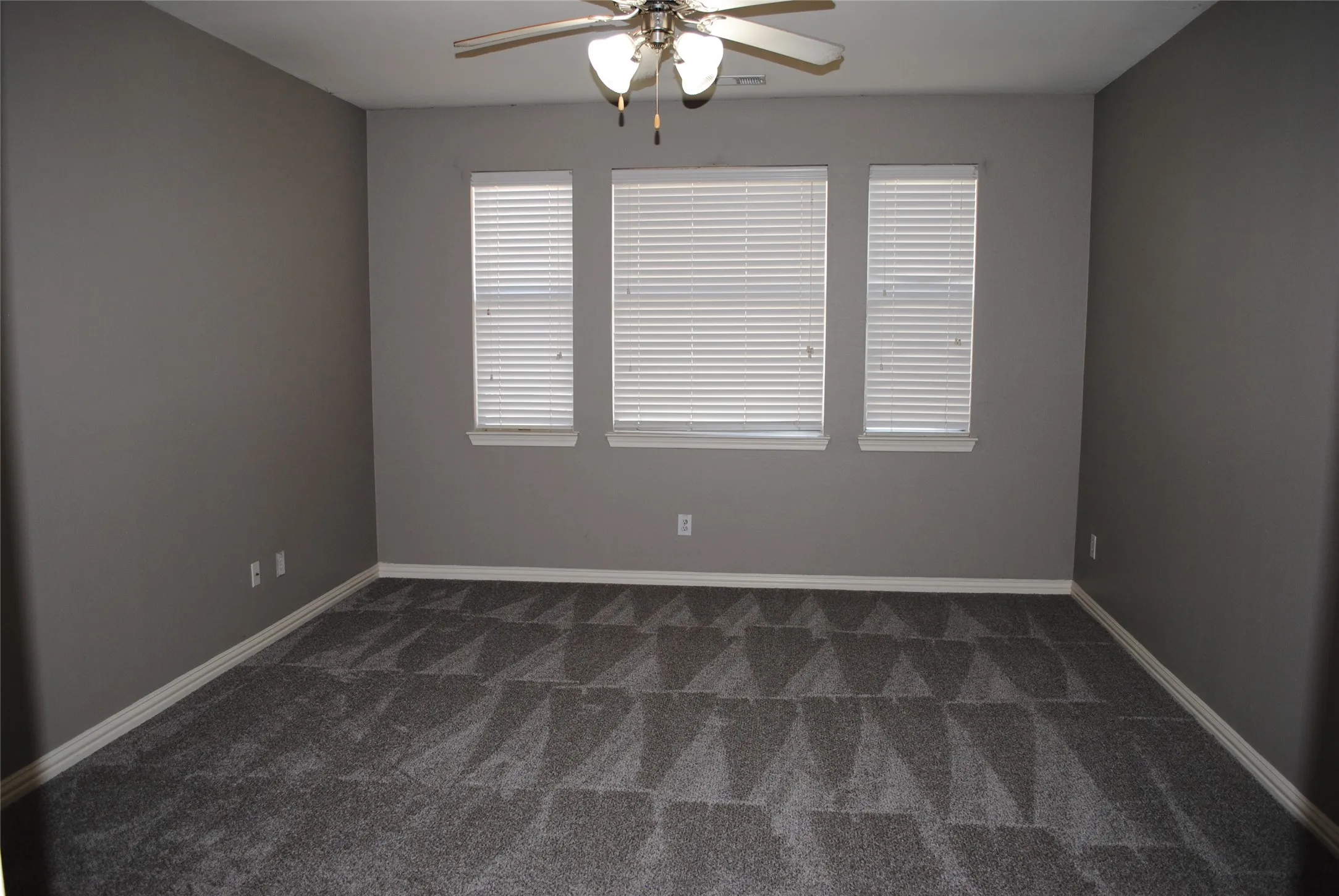Spacious room featuring neutral painted walls, three windows with blinds, and a ceiling fan with integrated lighting