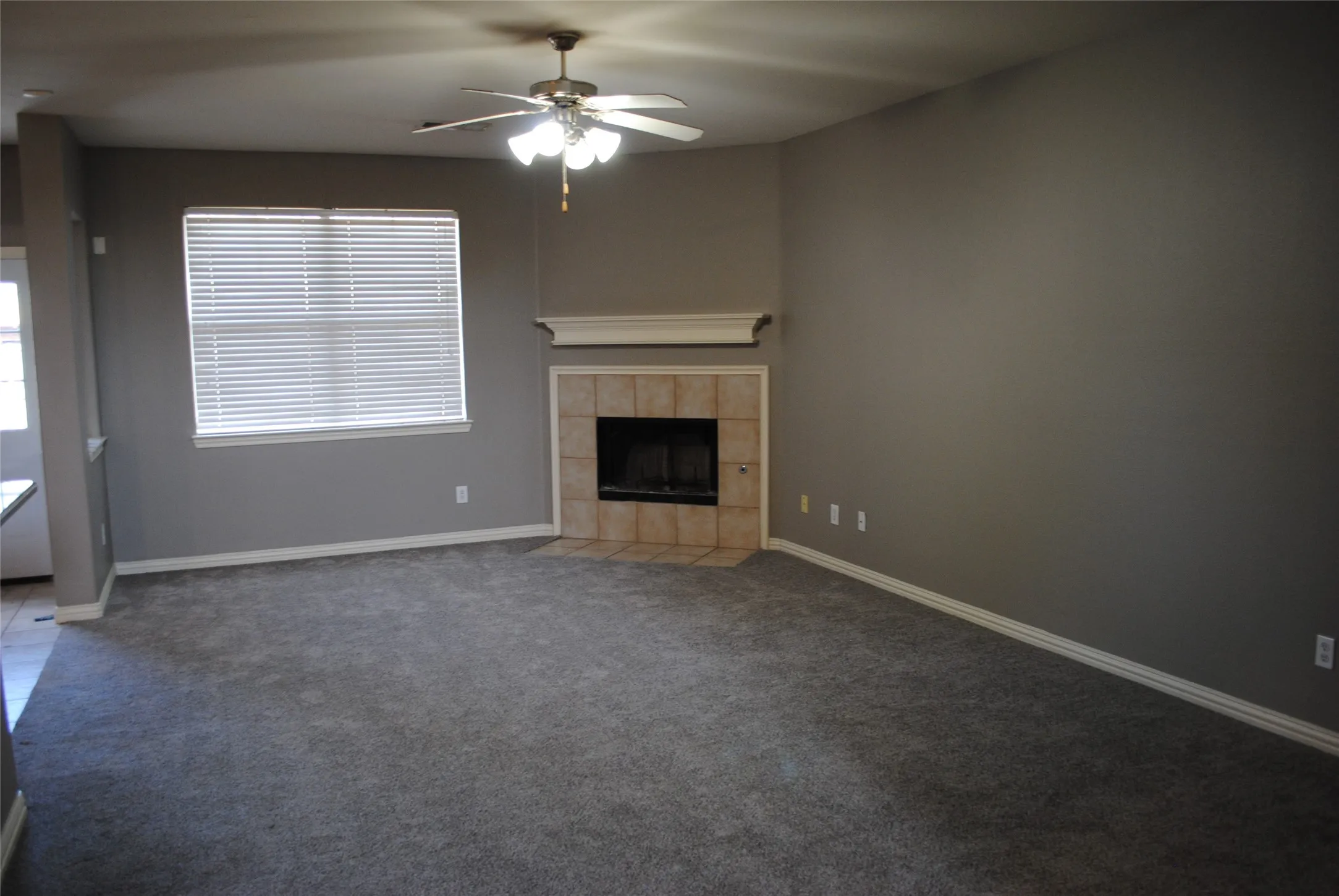 Spacious living area featuring plush carpeting, a corner fireplace with tile surround and mantel, and a ceiling fan with integrated lighting