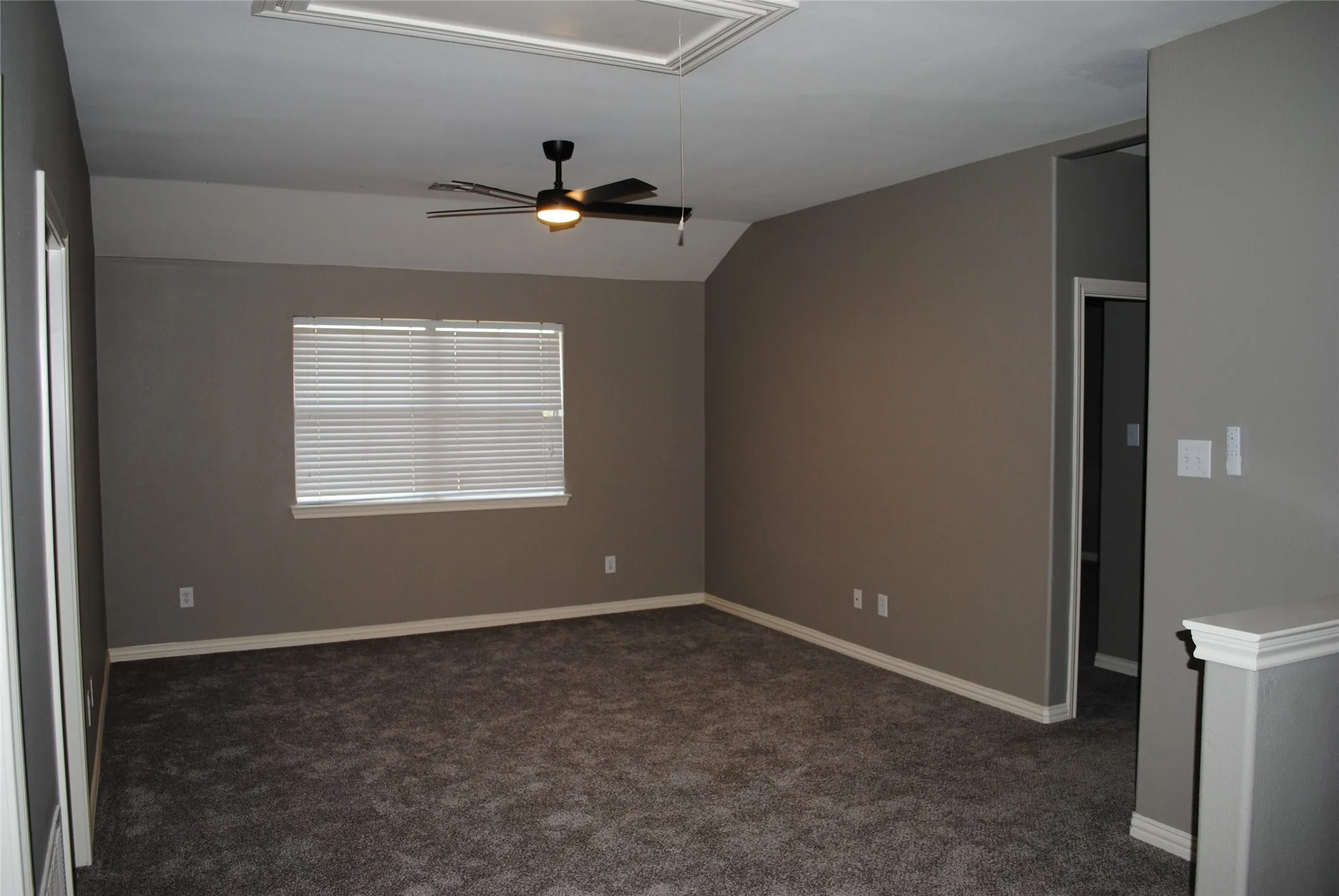 Spacious media room with gray wall paint, dark carpet flooring, window with horizontal blinds, and a ceiling fan with integrated lighting