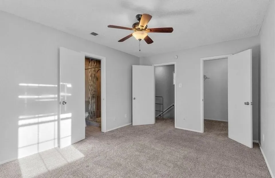A spacious primary bedroom showcasing an en suite bathroom, built-in cabinet, ceiling fan and neutral-colored carpeting, perfect for relaxation and comfort.