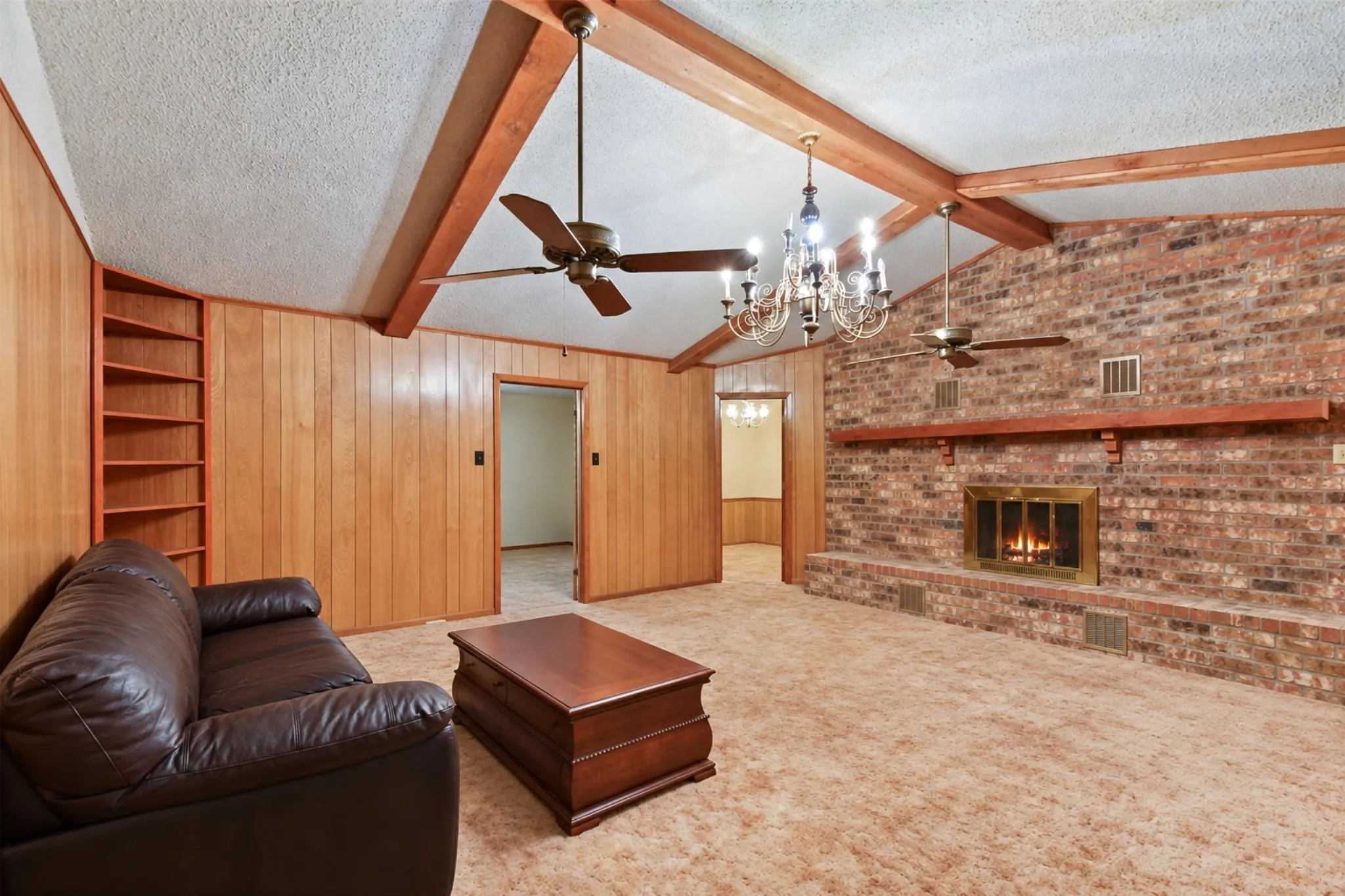 Brick fireplace with mantel, exposed wood beams, wood-paneled walls, built-in shelving, and ceiling fan