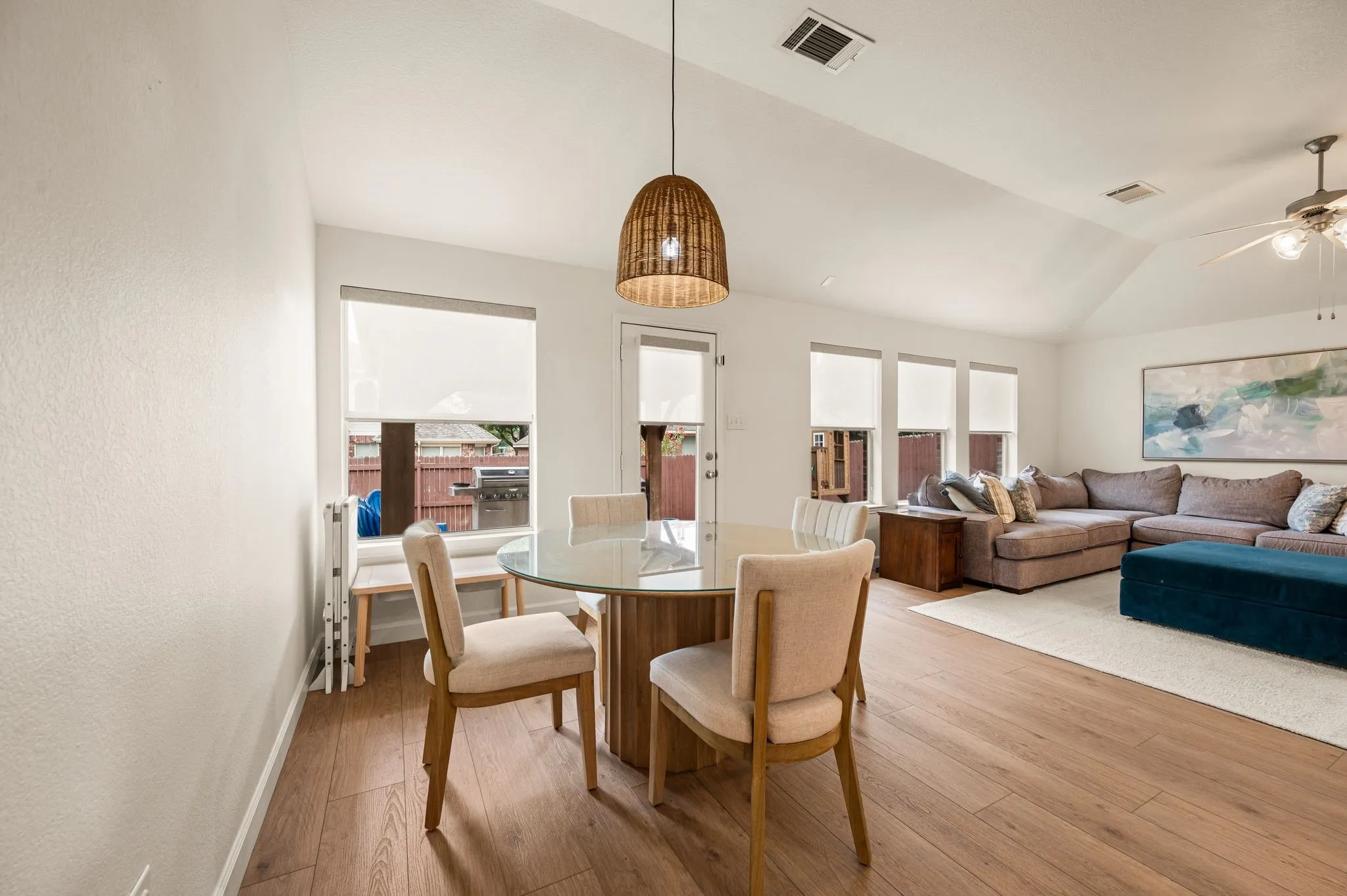 Vaulted ceiling with a ceiling fan, wood-finish flooring, multiple windows with roller shades, and a glass-top dining table with a basket-weave pendant light