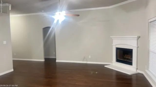 Wood-finish flooring, white crown molding, ceiling fan, fireplace with white mantel and hearth, and arched doorway
