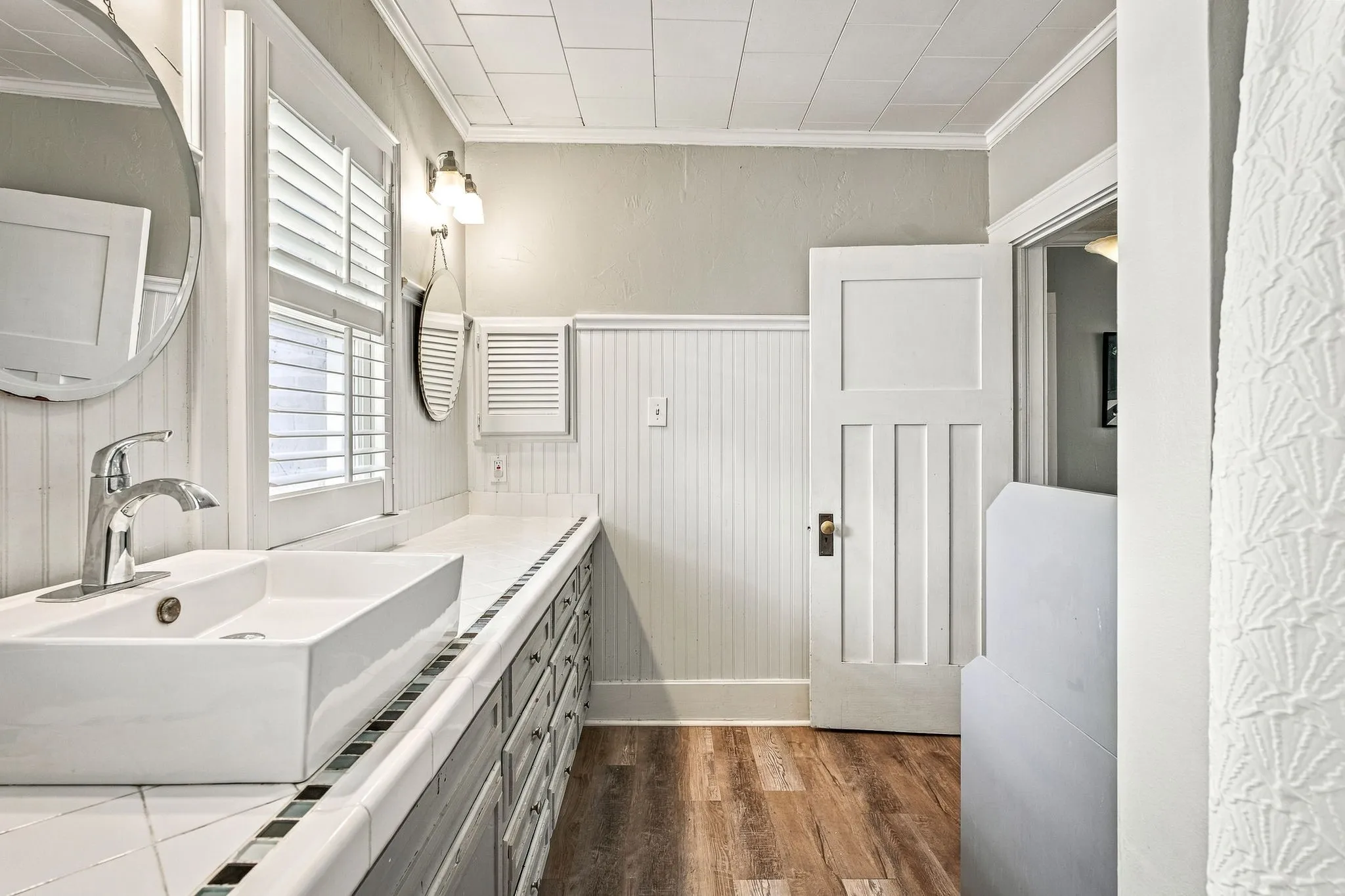 Bathroom featuring a vessel sink, wood-finish flooring, white beadboard wainscoting, white wooden shutters, and a paneled door