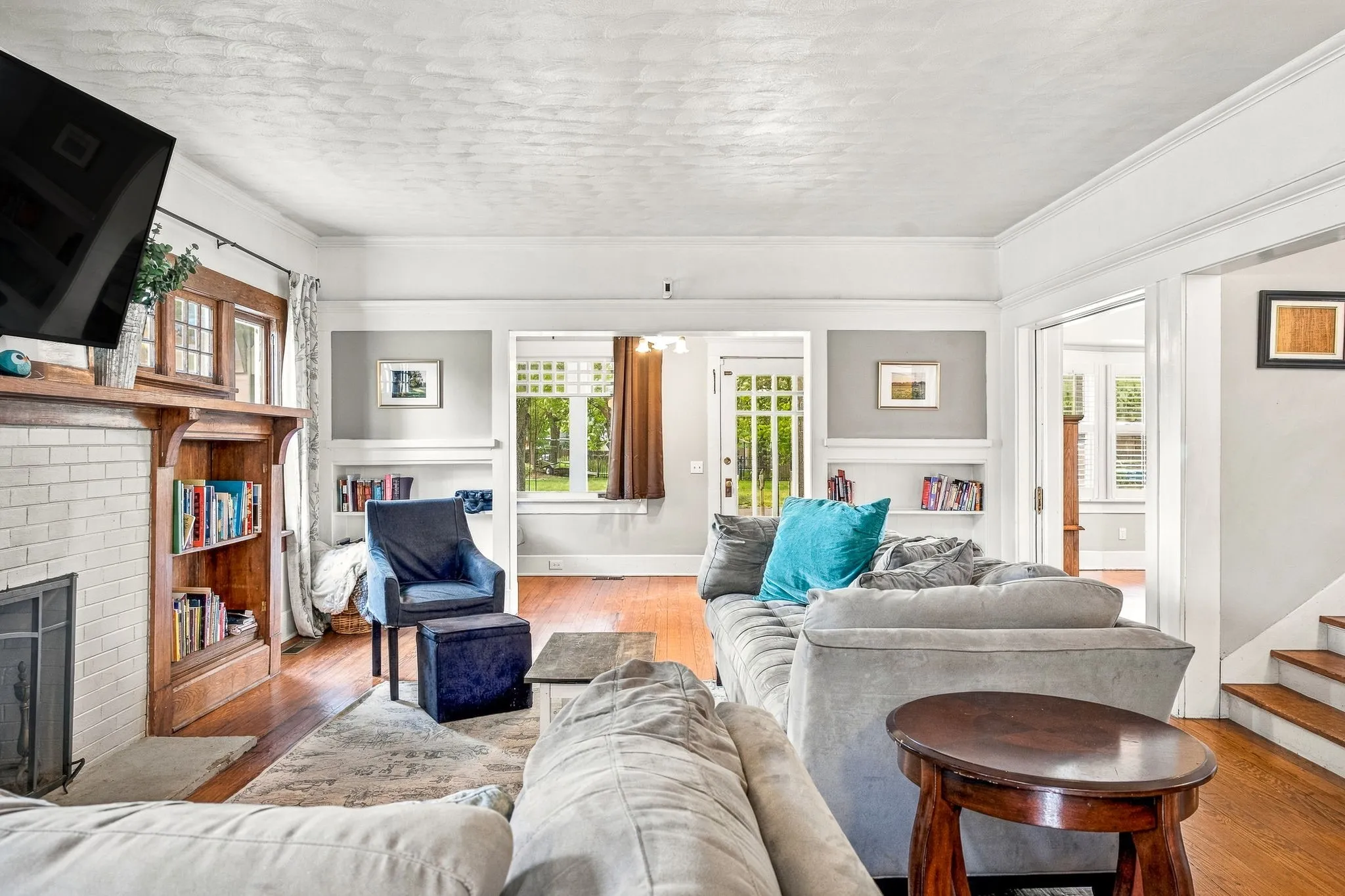 Hardwood flooring, built-in shelving, white brick fireplace, multiple windows, and crown molding