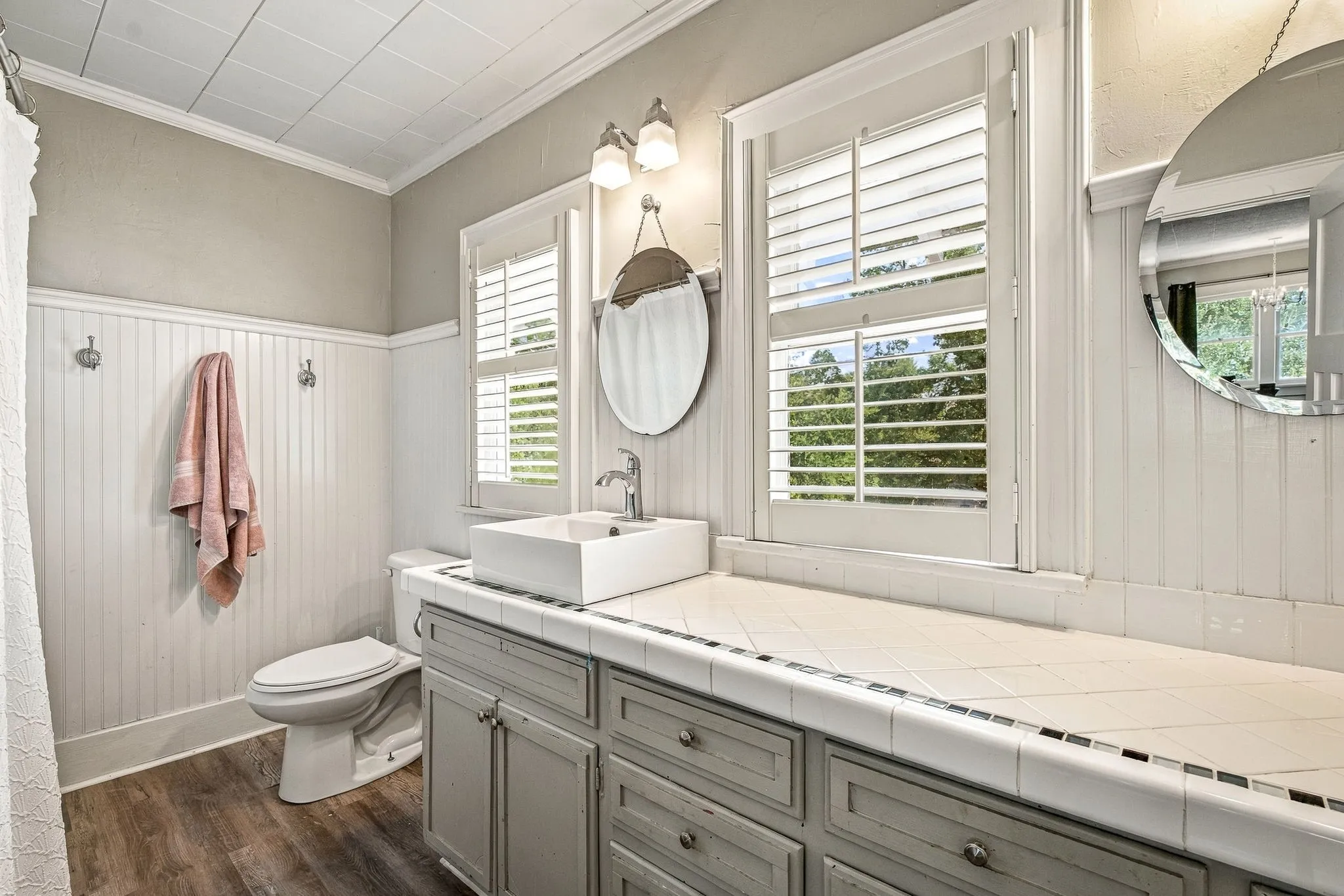 Wood-finish flooring, white beadboard wainscoting, and a white tiled countertop with a vessel sink