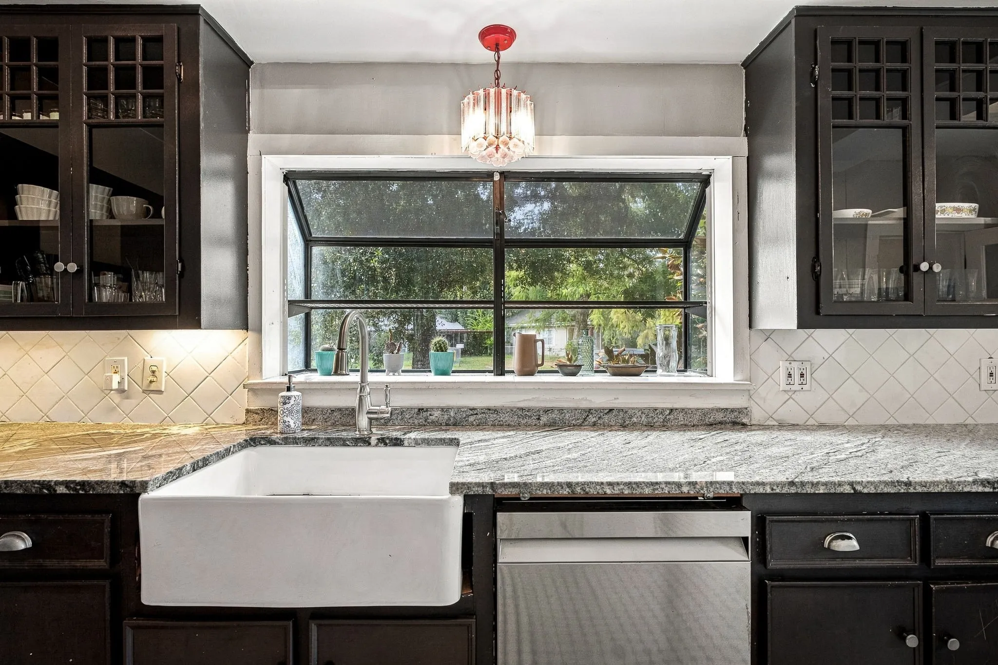 Kitchen featuring an apron-front sink, granite countertops, dark cabinetry with glass inserts, a stainless steel dishwasher, and a casement window