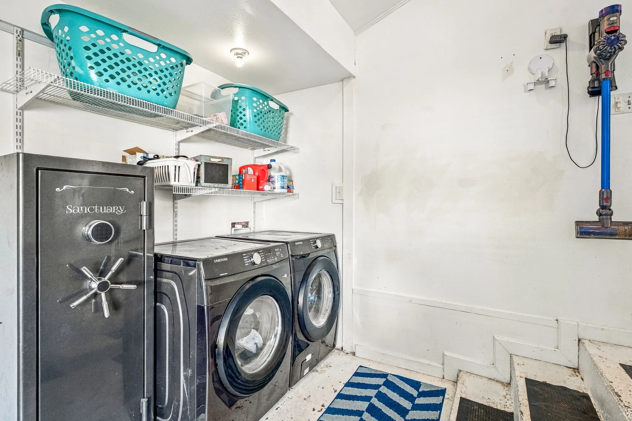 Dedicated laundry room featuring side-by-side front-load washers and dryer, open shelving with turquoise storage baskets, granite countertop workspace, and neutral tile flooring with striped accent mat