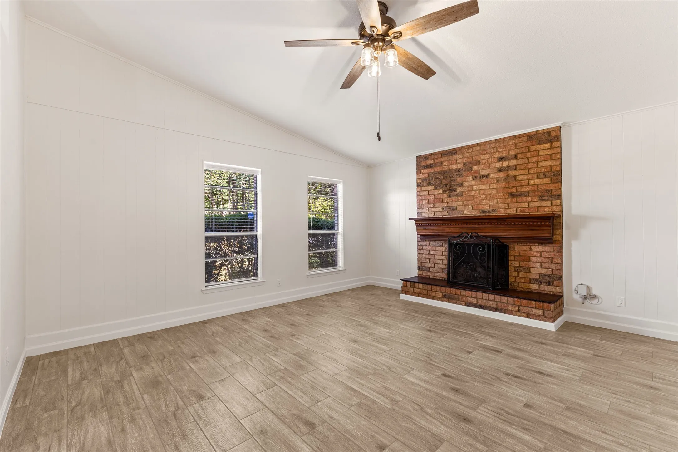 Vaulted ceiling, wood-finish flooring, brick fireplace with decorative mantel, ceiling fan with integrated lighting, and white paneled walls
