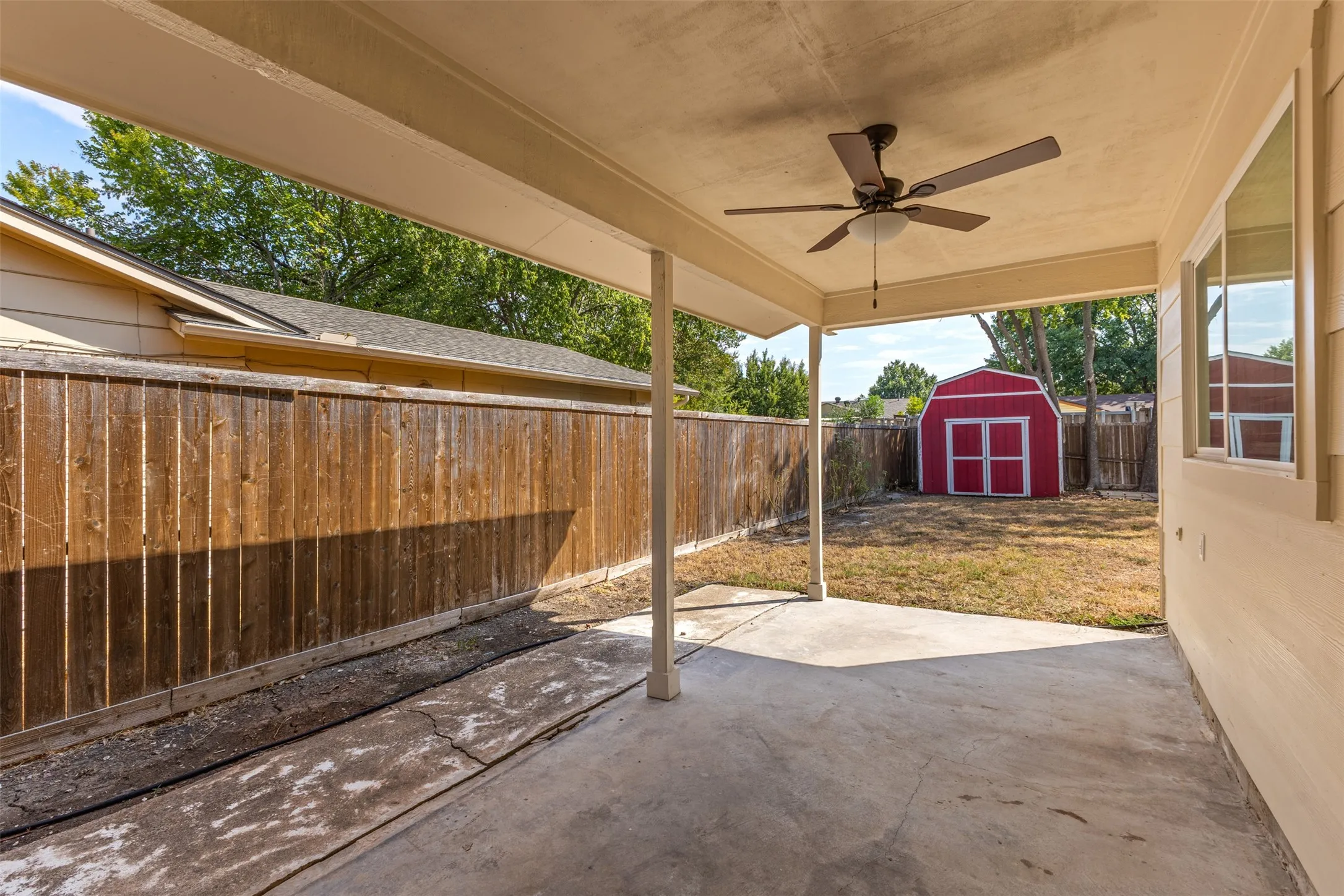 Covered patio with concrete flooring, supported by structural pillars, featuring an attached ceiling fan