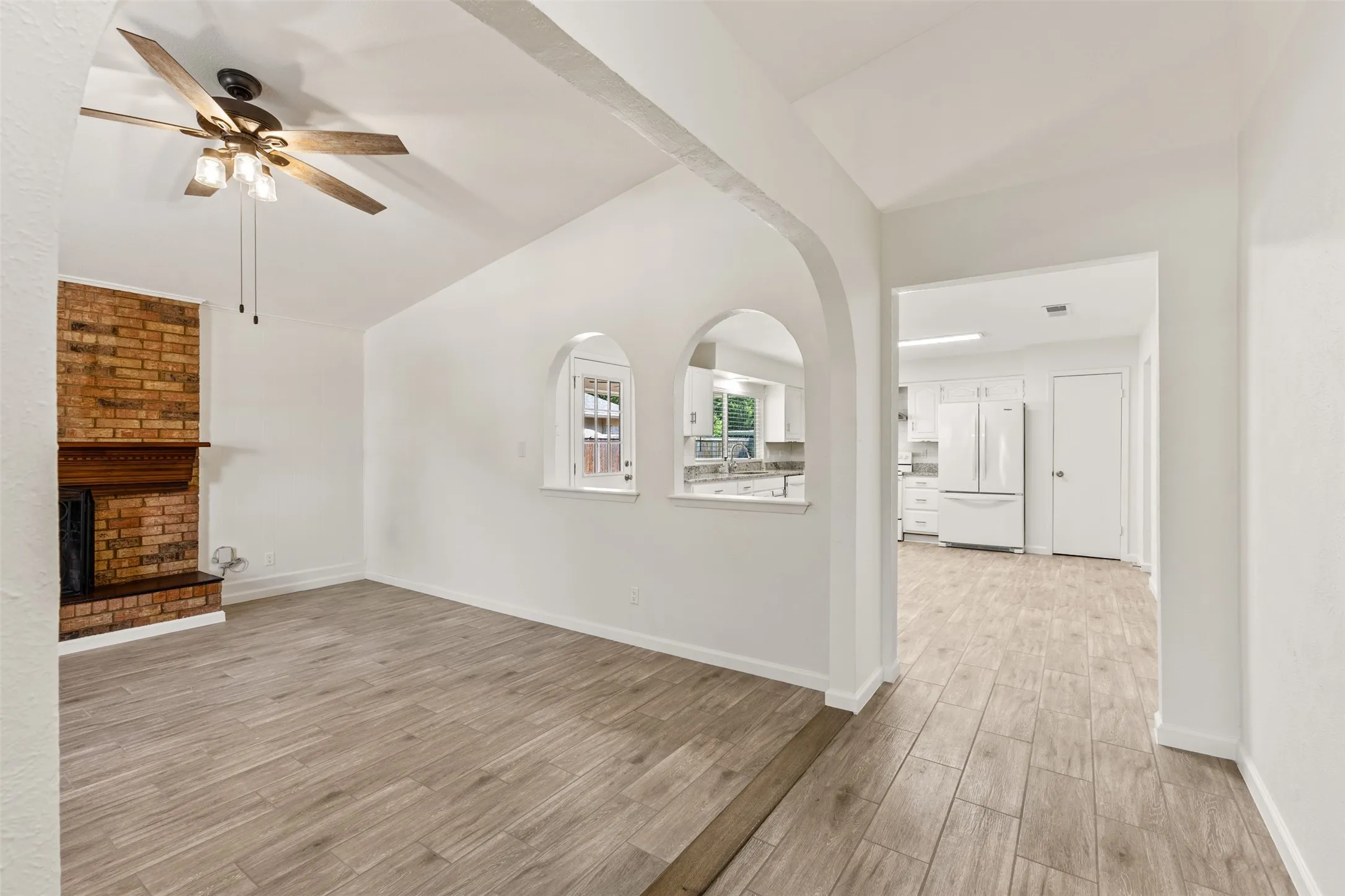 Vaulted ceiling with a ceiling fan, brick fireplace with wood mantel, arched wall openings, and wood-finish flooring