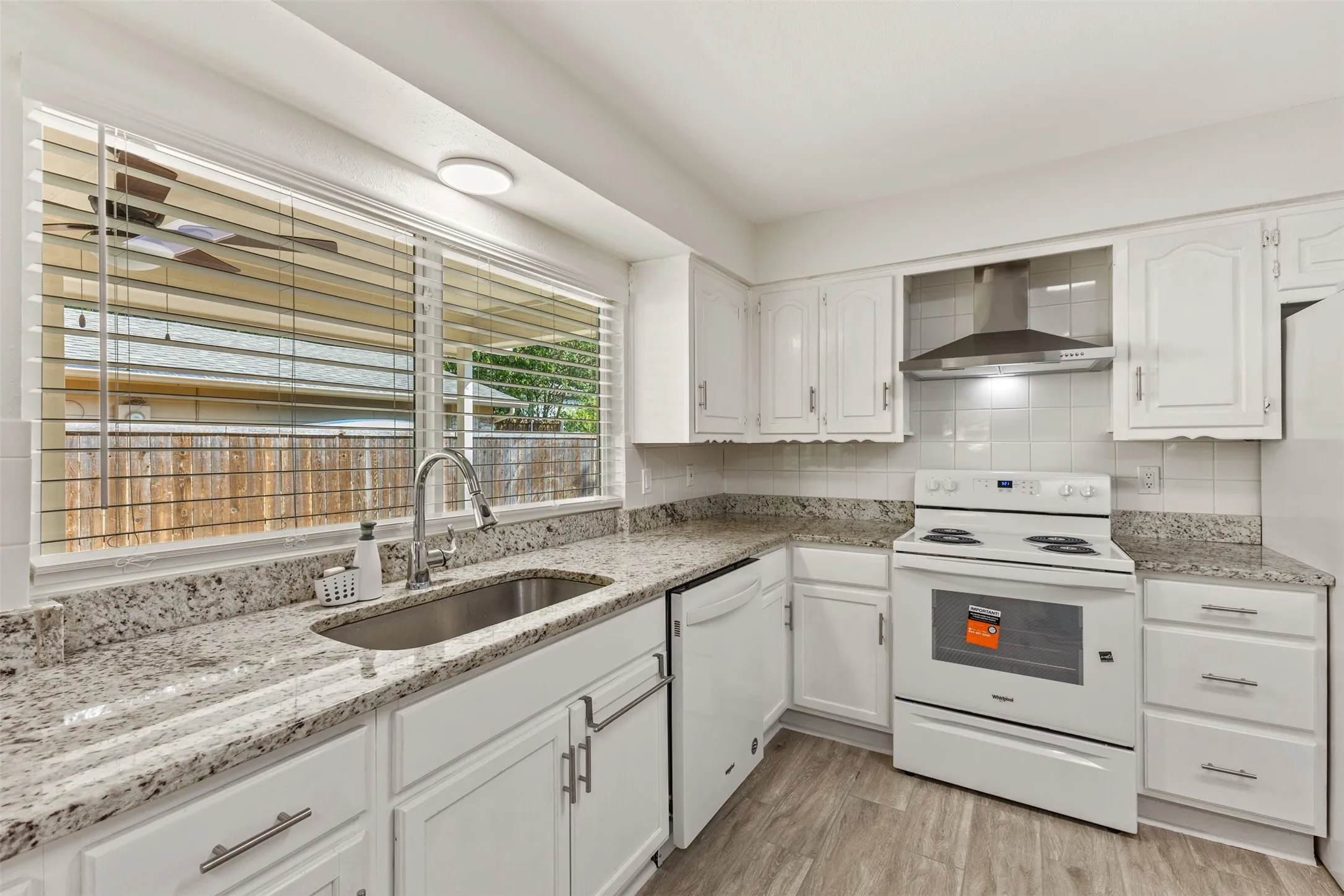 White cabinetry, speckled countertops, stainless steel sink, wood-finish flooring, and a stainless steel range hood
