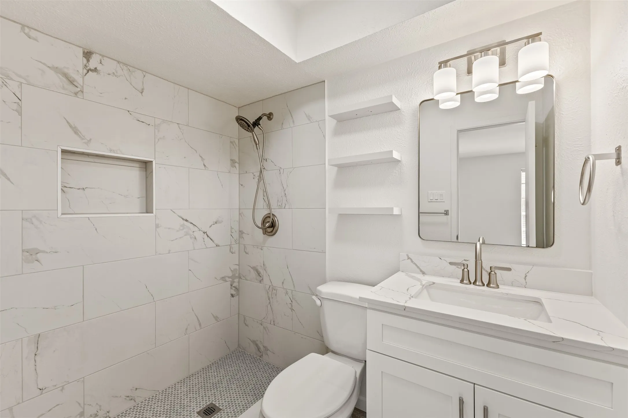 Full tile shower surround with integrated niche, mosaic tile shower pan, brushed nickel shower fixture, white vanity with integrated sink and quartz countertop, and vanity lighting fixture