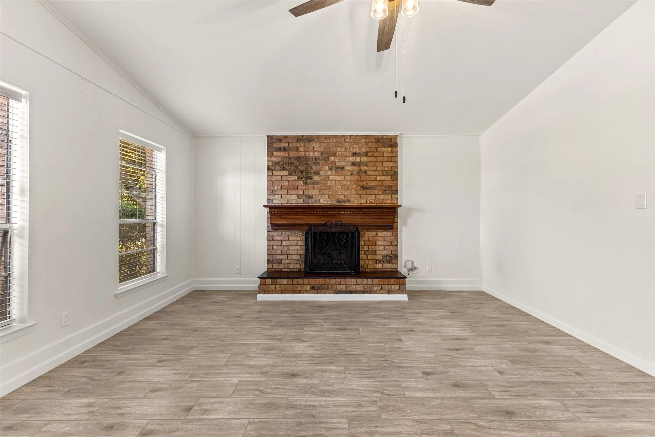 Spacious living area featuring a brick fireplace with a wood mantel, wood-finish flooring, vaulted ceiling, and a ceiling fan with light fixture