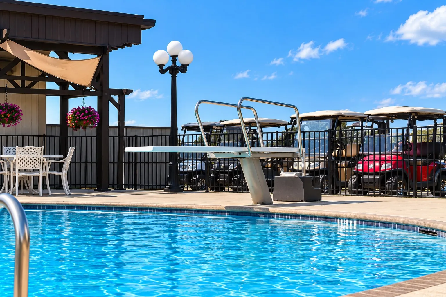 Resort-style pool with diving board, surrounded by a paved deck, permanent lamp post, and a black wrought-iron fence