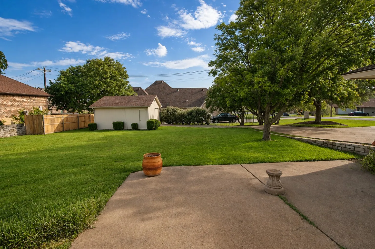Expansive lawn with mature trees, concrete patio, stone retaining wall, and a wood privacy fence