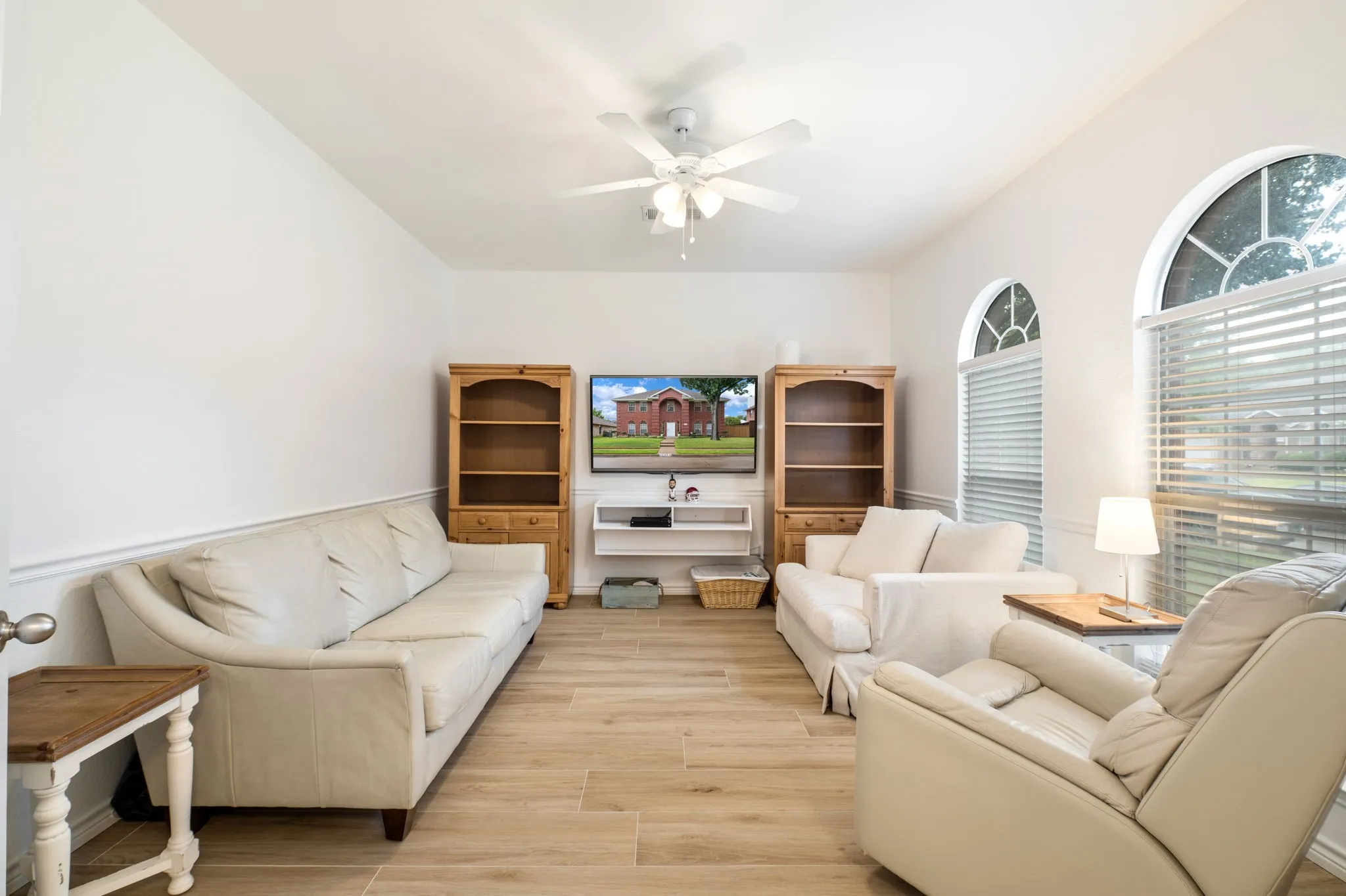 Bright, open living room featuring hardwood flooring, ceiling fan with integrated lighting, built-in wooden shelving units flanking an entertainment center, and large windows with plantation shutters providing natural illumination and privacy control