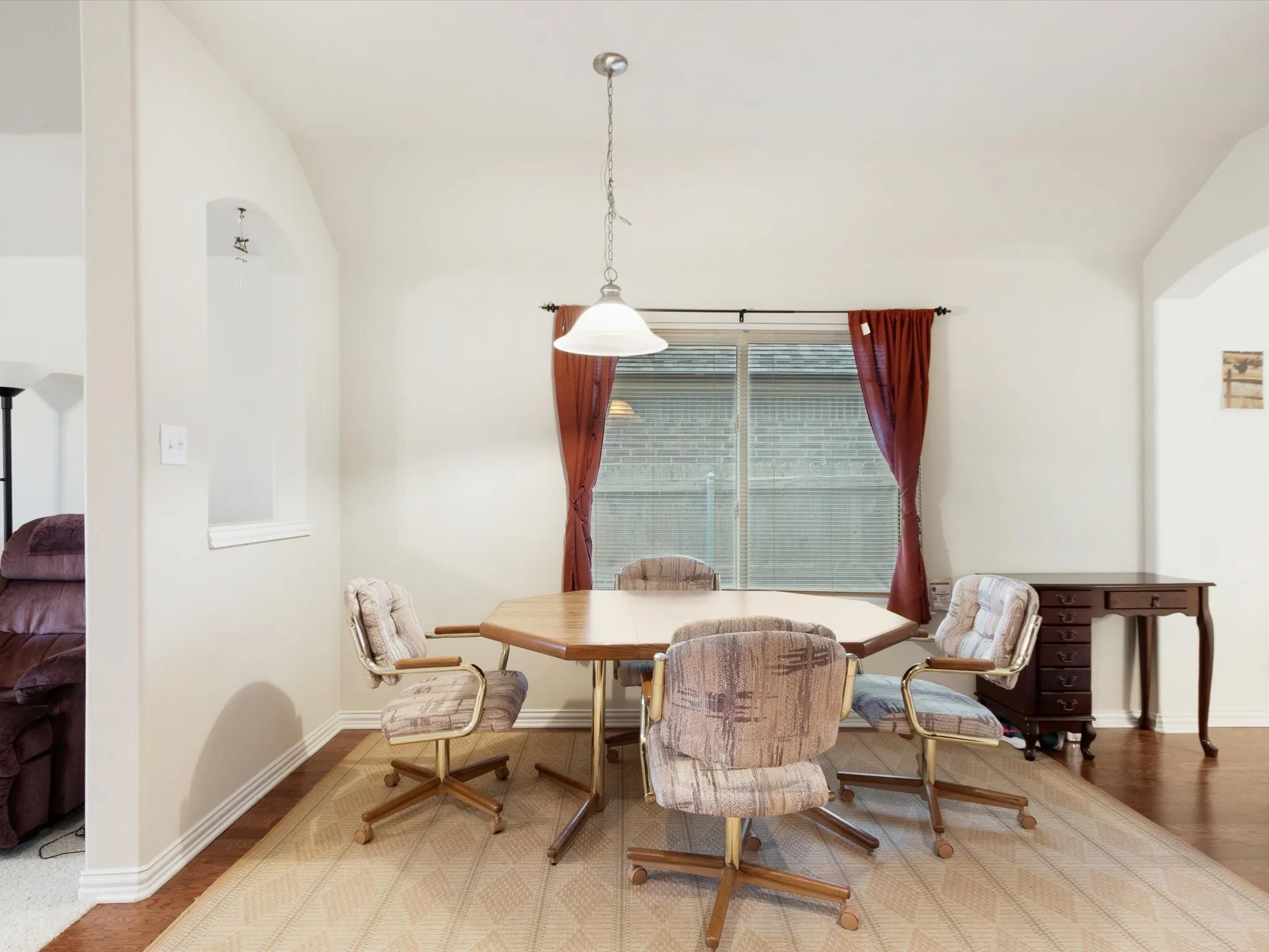 Arched wall opening, window with horizontal blinds, overhead pendant light fixture, and wood-finish flooring