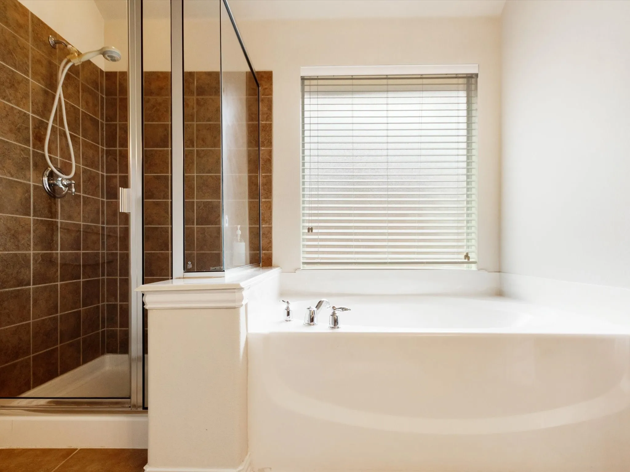 Built-in soaking tub with deck-mounted fixtures, separate shower with tiled walls and glass enclosure, and a window with blinds