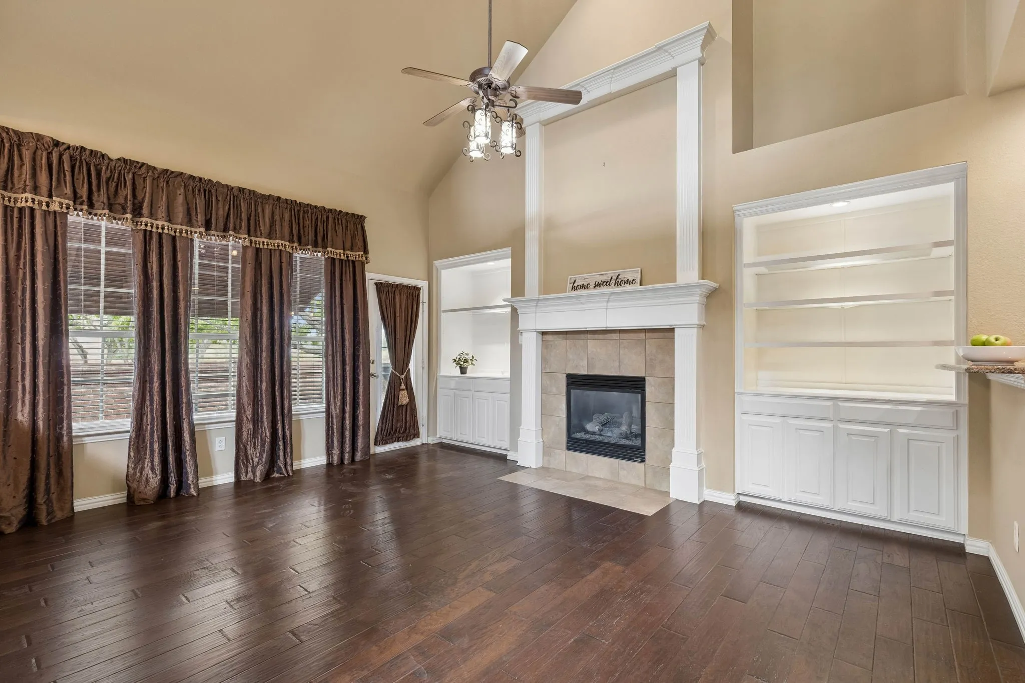 Vaulted ceiling with a ceiling fan, dark wood-finish flooring, built-in shelving, gaslog fireplace with a tile surround, and multiple windows looking over the patio
