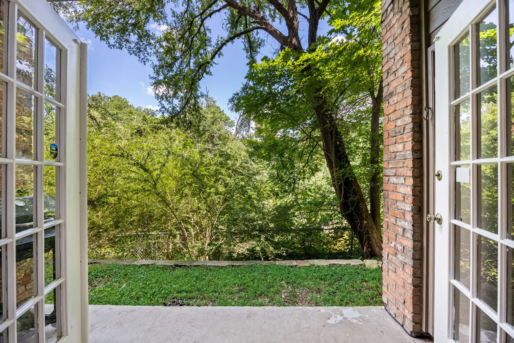 Patio with concrete flooring, brick exterior wall, and double French doors opening to a grassy area and mature trees