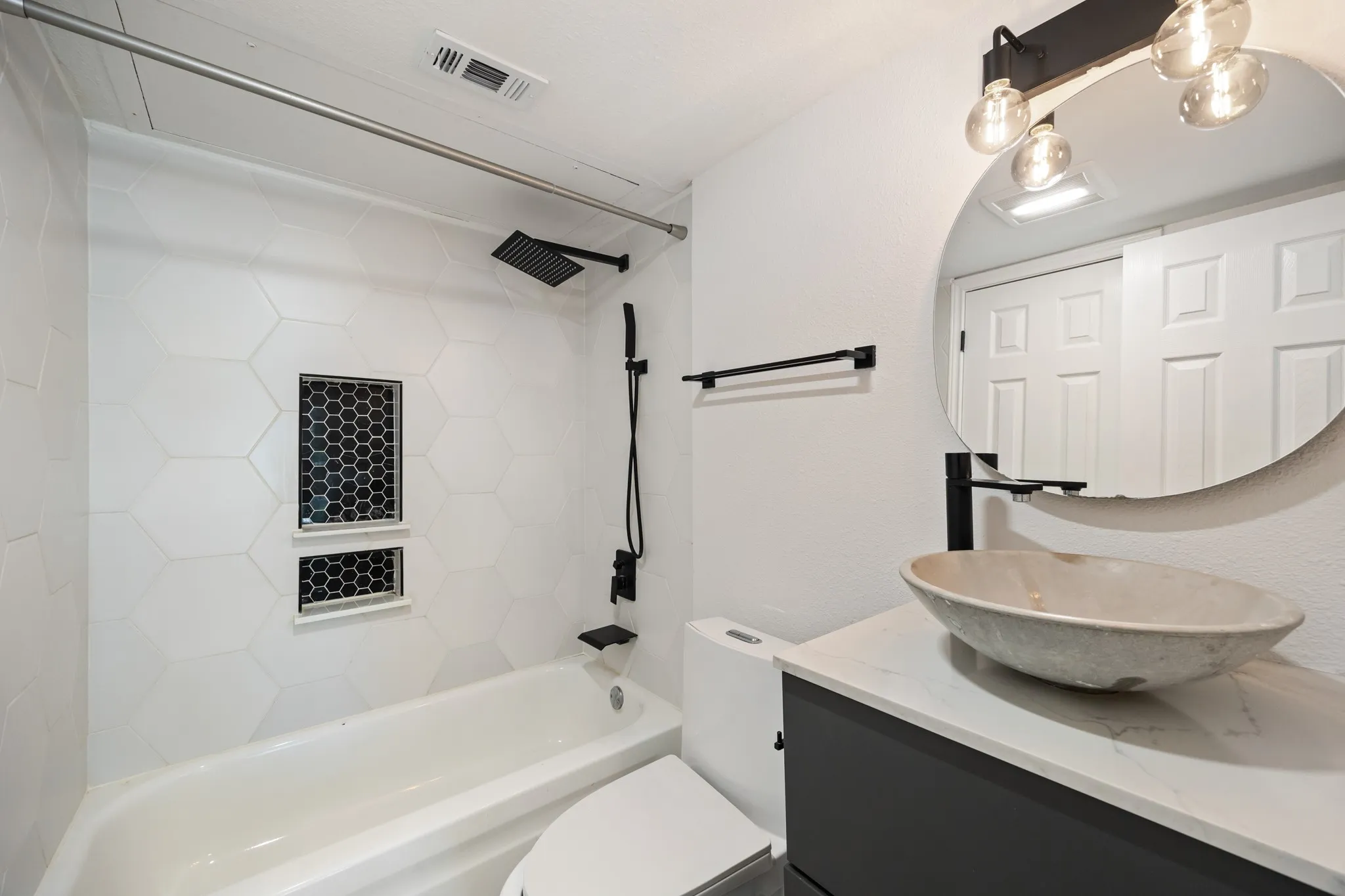 White hexagonal wall tile, dual shower niches with mosaic tile, black matte rainfall showerhead, vessel sink, and a black vanity with a white countertop