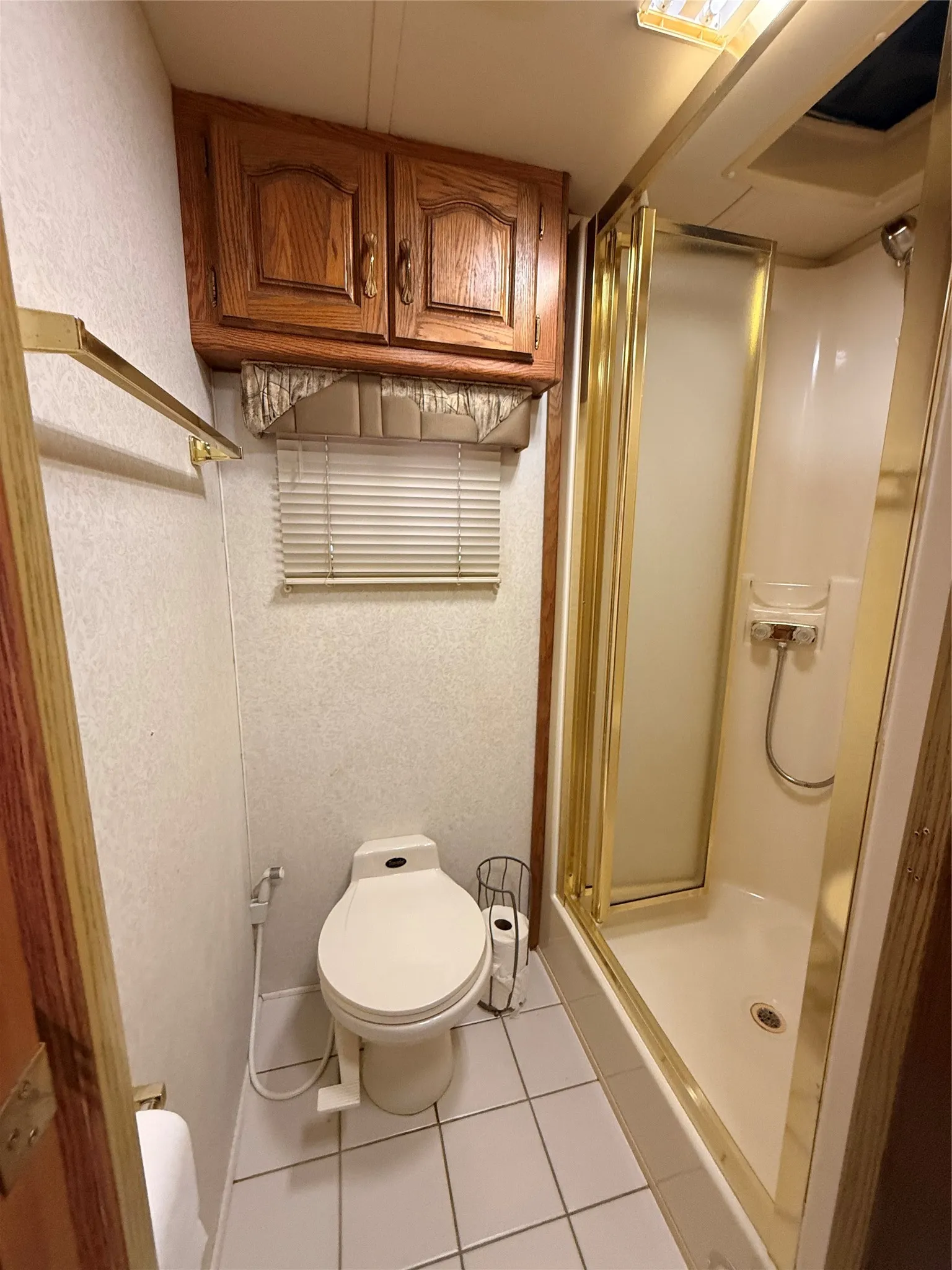 Tiled flooring, overhead wood-finish cabinetry, shower enclosure with a gold-tone frame, and window with blinds
