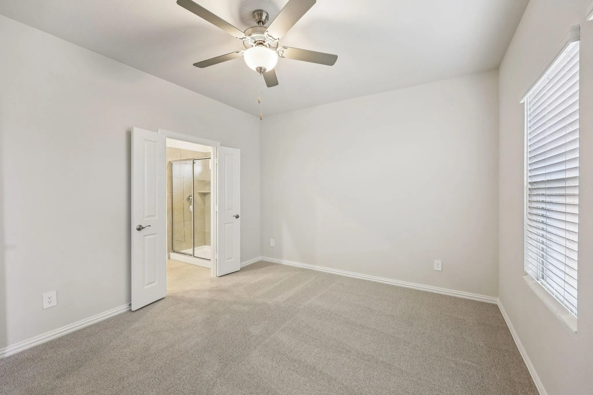 Neutral wall paint, light-toned carpeting, ceiling fan with light fixture, window with blinds, and double doors leading to a separate room with a glass-enclosed shower