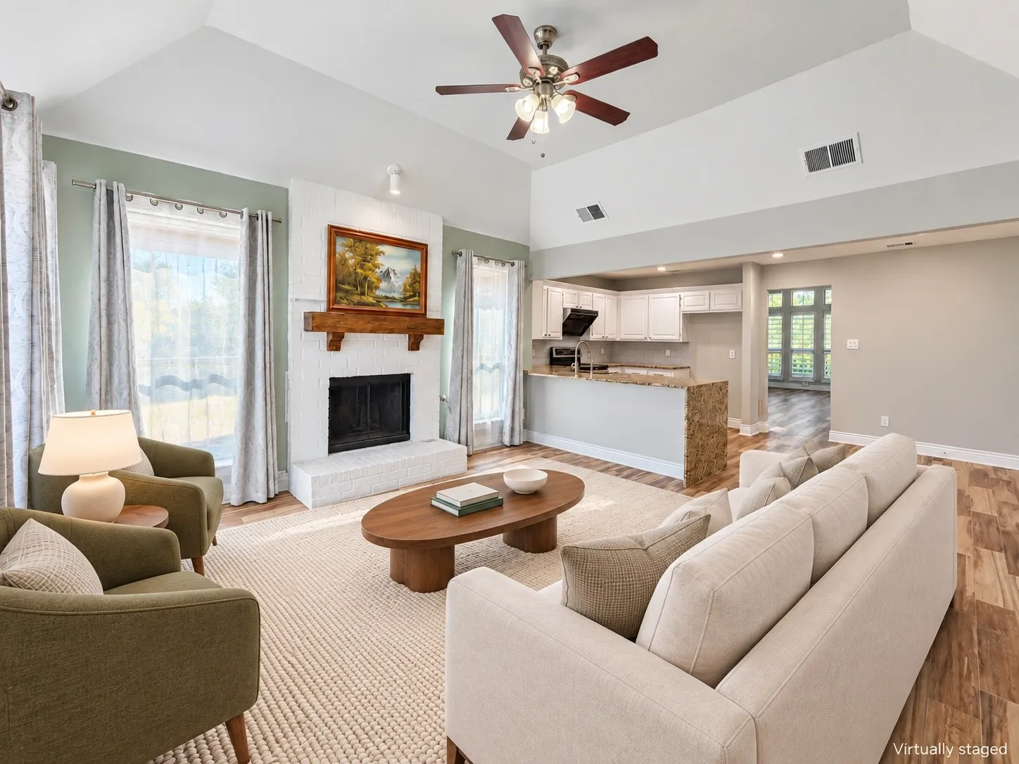 Vaulted ceilings with a ceiling fan, a white brick fireplace with a wood mantel, wood-finish flooring, and a kitchen pass-through with a stone-finish countertop