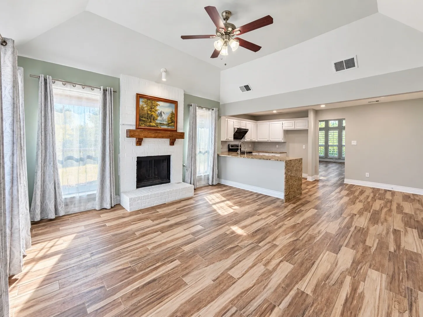 Vaulted ceiling, wood-finish flooring, white brick fireplace with wood mantel, ceiling fan, and a kitchen pass-through with a stone-finish countertop