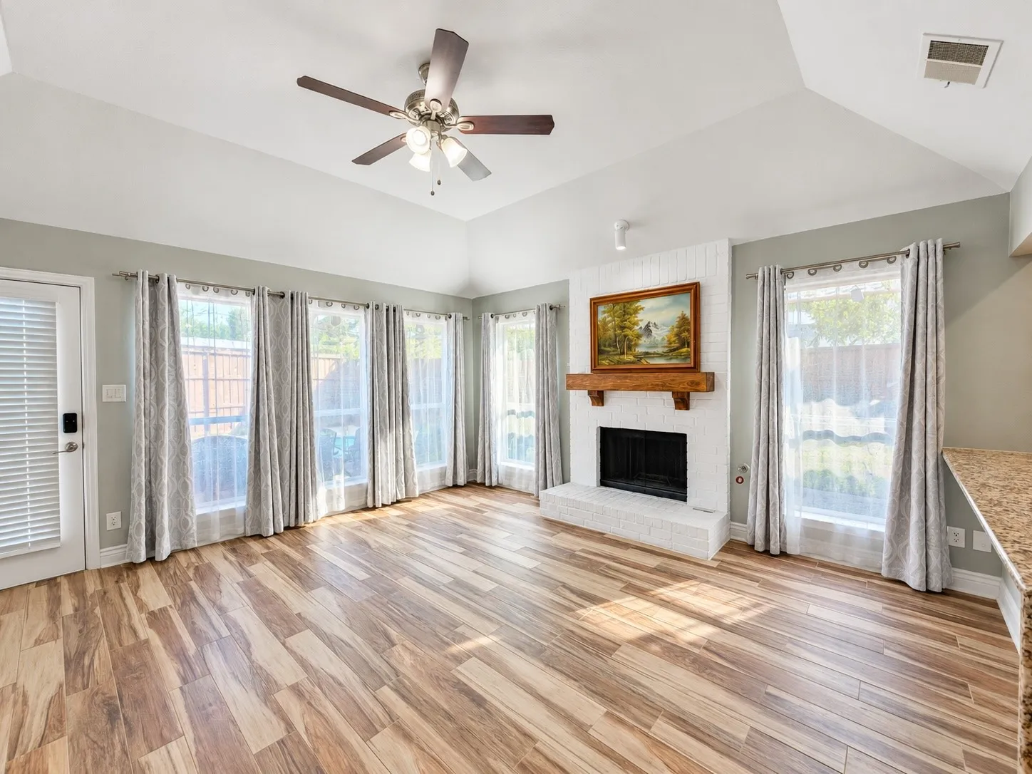 Vaulted ceiling with a ceiling fan, brick fireplace with a wood mantle, wood-finish flooring, and multiple windows
