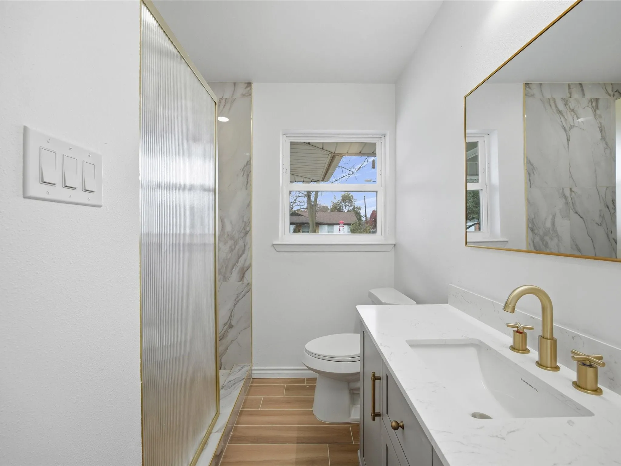 Walk-in shower with opaque glass paneling, wood-finish flooring, single vanity with integrated sink, rectangular gold-framed mirror, and gold-finish fixtures