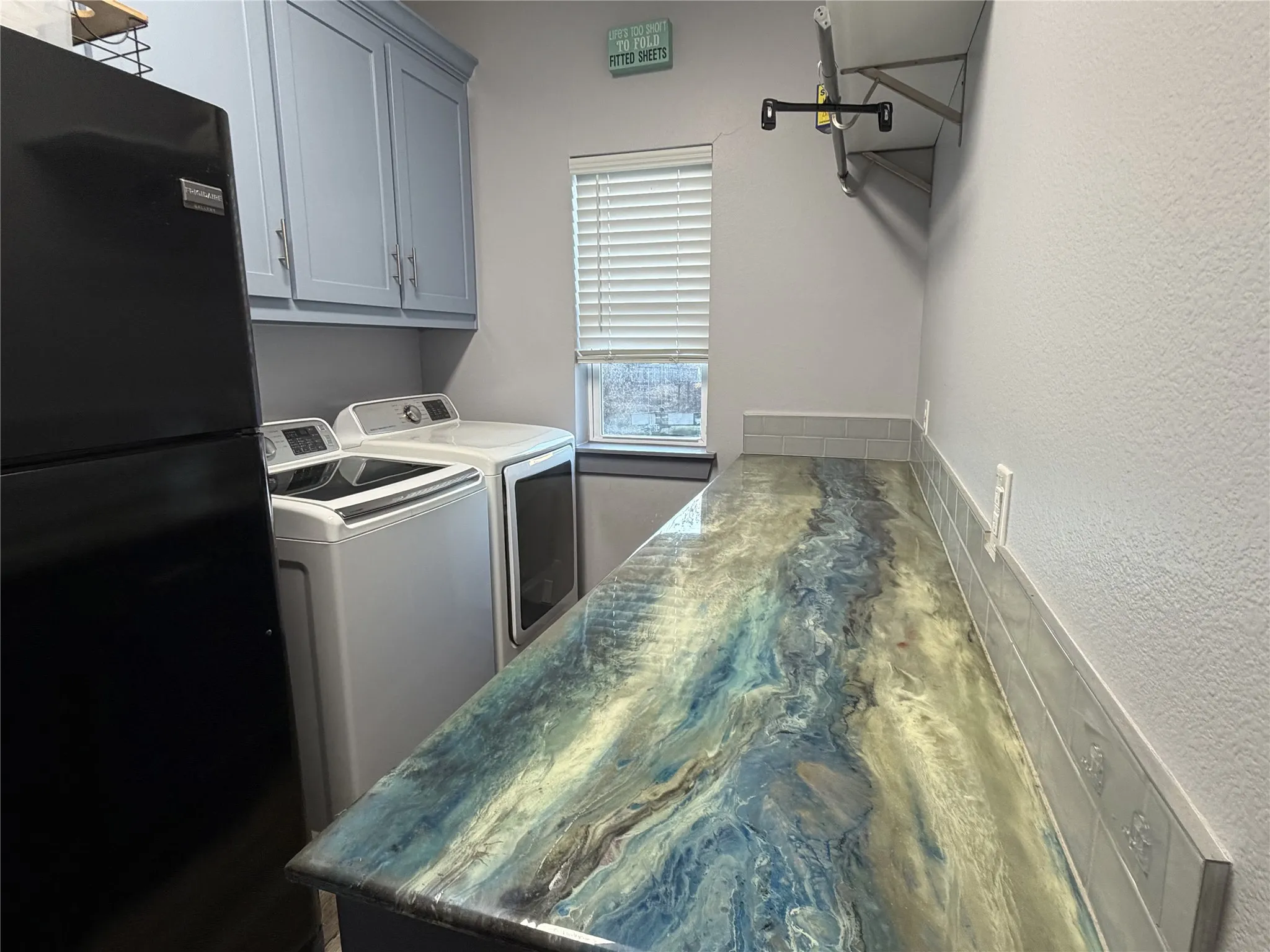 Custom countertop finish, subway tile backsplash, window with horizontal blinds, overhead cabinetry, and wall-mounted shelving