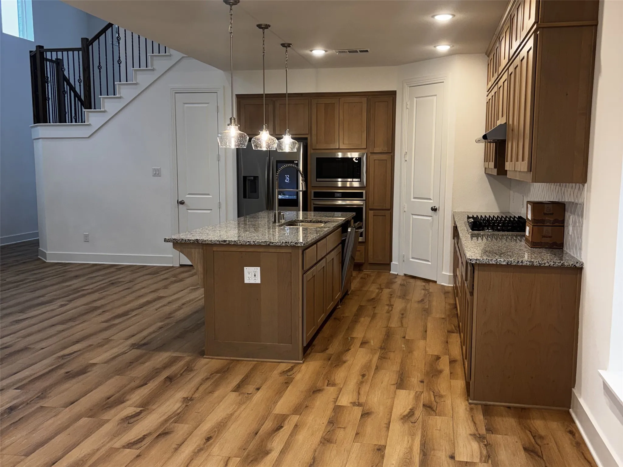 Modern kitchen featuring stainless steel appliances, wood-finish flooring, granite countertops, a central island with sink, and pendant lighting