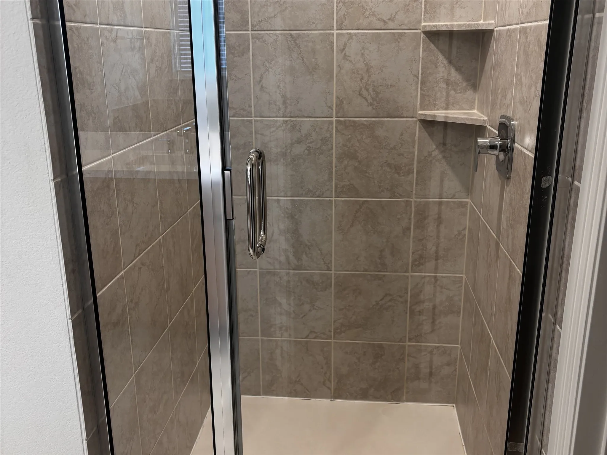 Enclosed shower with tiled walls, integrated corner shelving, chrome fixtures, and a clear glass door with a polished handle